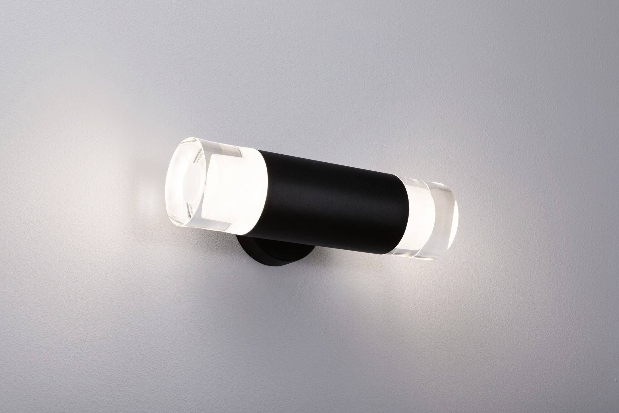 Eine moderne Wandleuchte mit zylindrischer Form, schwarzem Mittelteil und transparenten Enden erleuchtet eine weiße Wand.