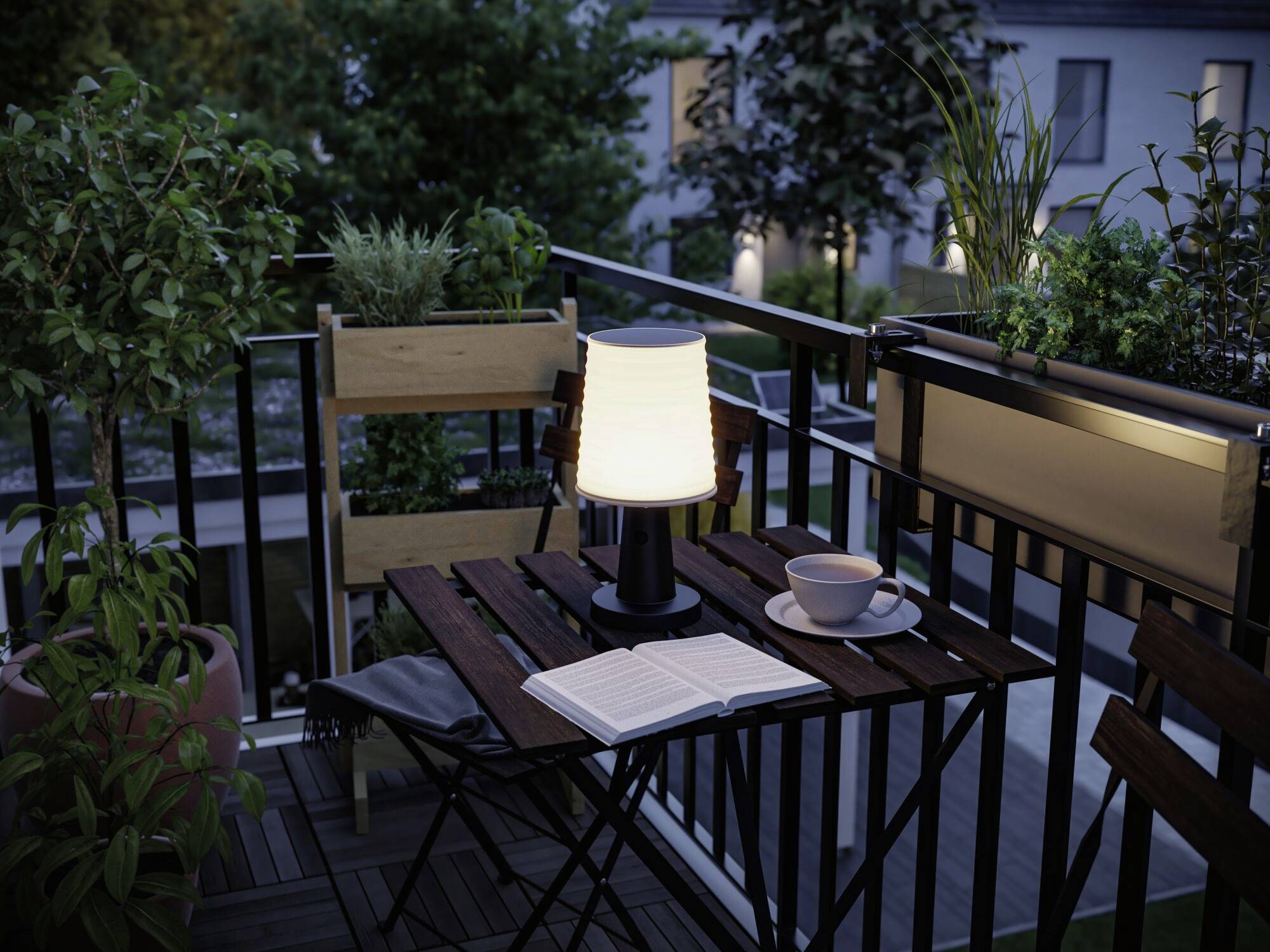 Der Balkon zeigt einen Tisch mit einer Lampe, einem geöffneten Buch und einer Tasse, umgeben von Pflanzen und Abendbeleuchtung.