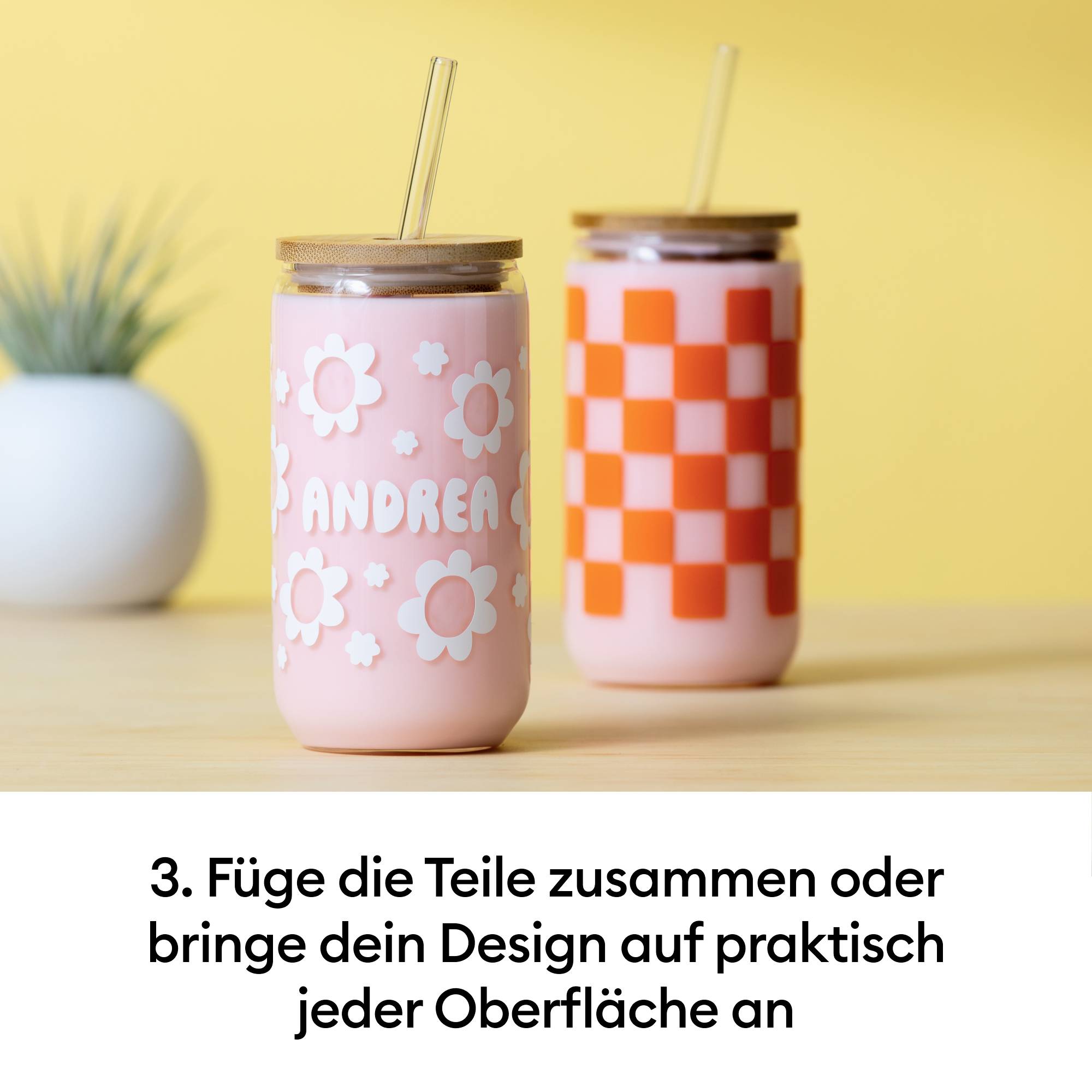 Zwei pastellfarbene Trinkbecher mit Bambusdeckeln und Strohhalmen. Auf einem Becher steht 'Andrea'. Hintergrund ist unscharf.