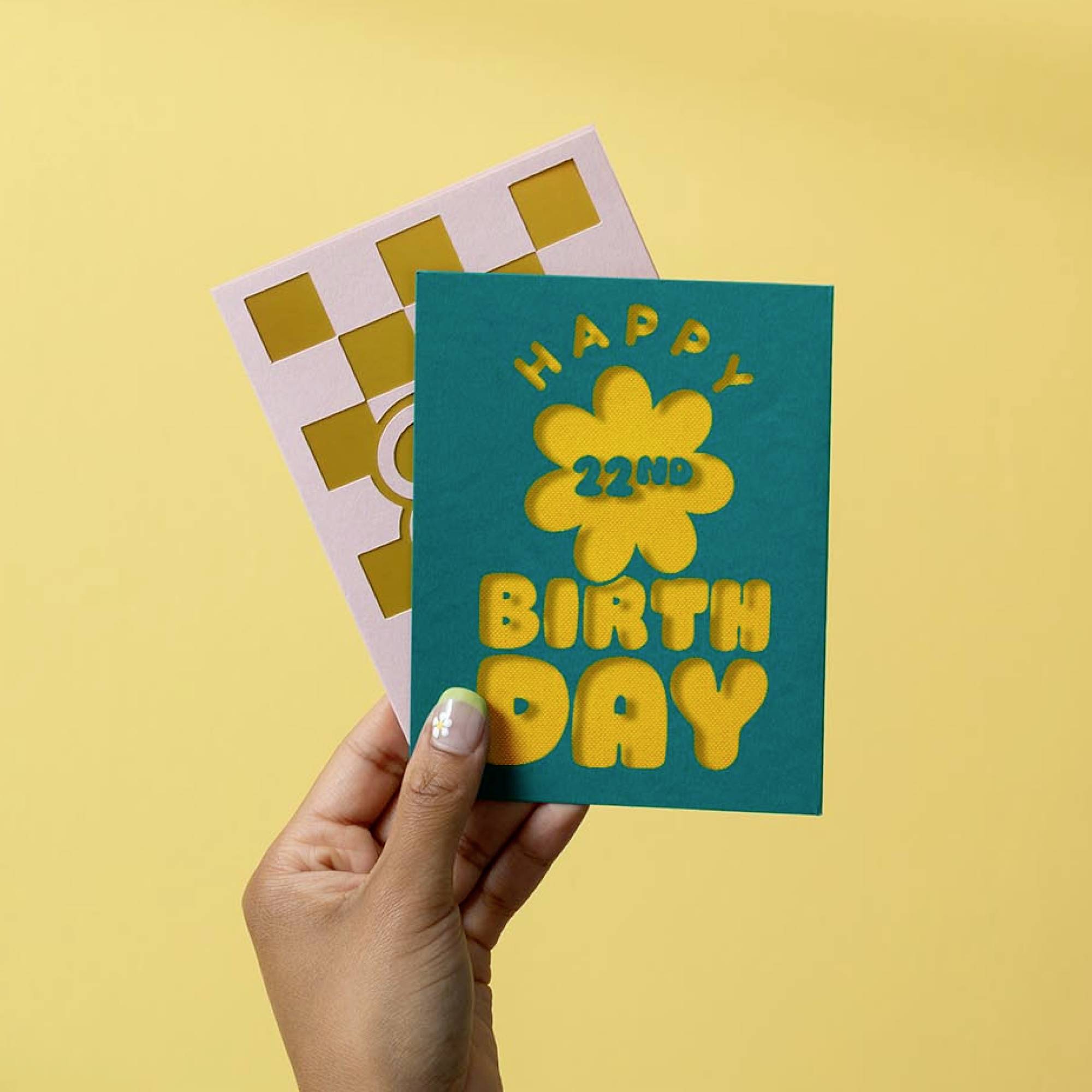 Eine Hand hält zwei Grußkarten vor gelbem Hintergrund. Die vordere Karte zeigt die Aufschrift 'Happy 22nd Birthday' mit Blumenmotiv.