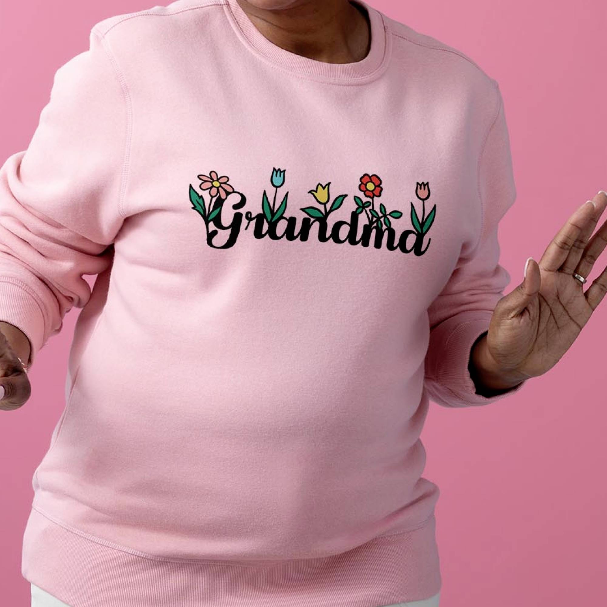 Eine Person trägt einen rosa Pullover mit der Aufschrift 'Grandma' und floralen Motiven vor einem rosa Hintergrund.