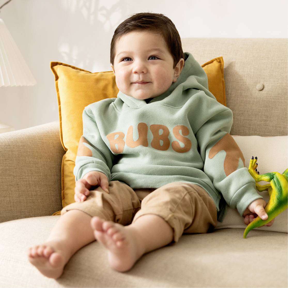 Kleinkind in grünem Hoodie sitzt auf einem Sofa, umgeben von gelben Kissen. Lächelt und schaut in die Kamera.