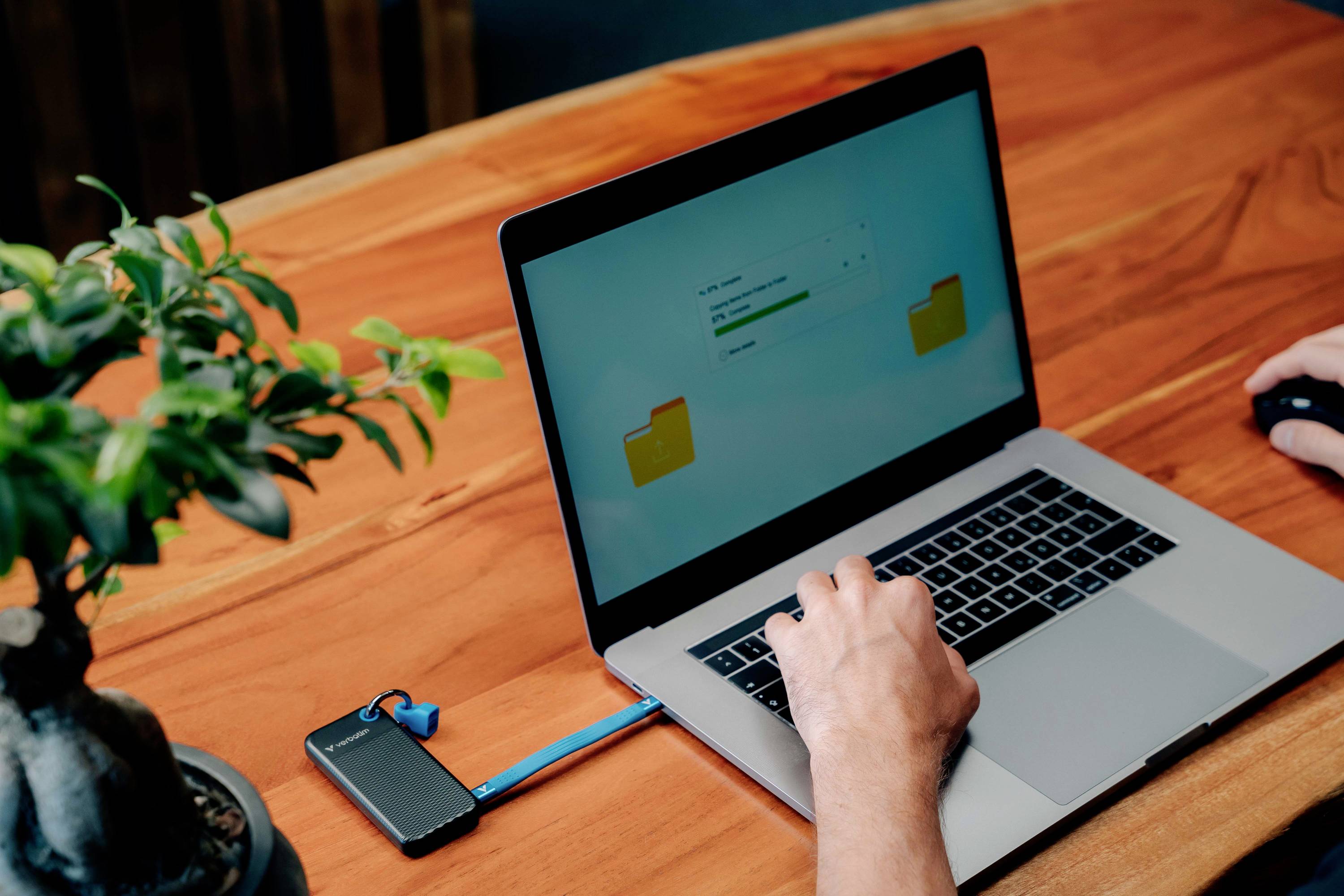 Laptop auf Holztisch mit angeschlossenem tragbaren Laufwerk, das Daten überträgt; eine Person benutzt die Maus. Büropflanze daneben.
