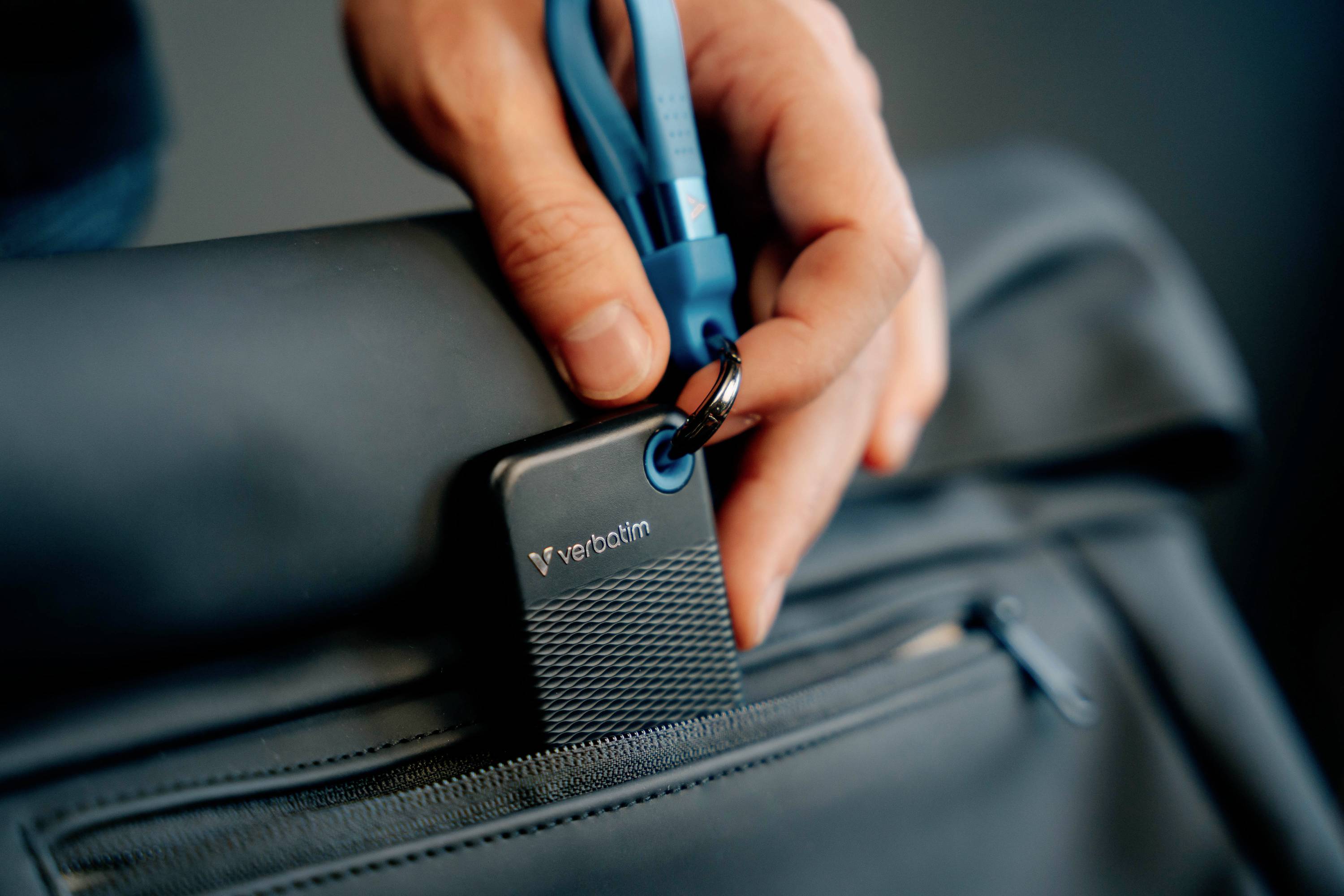 Eine Hand befestigt eine tragbare SSD-Festplatte mit einem blauen Kabel an einem schwarzen Rucksack.