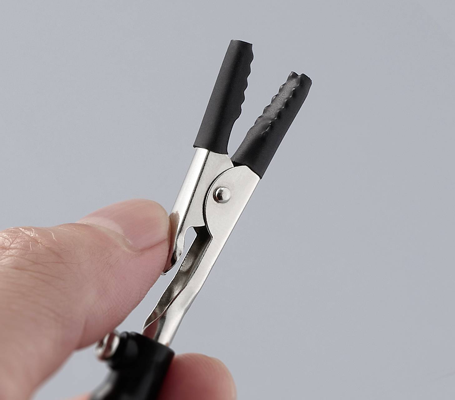 Eine Hand hält eine kleine Metallklemme mit schwarzen Gummispitzen. Die Klemme ist geöffnet und wird gegen einen grauen Hintergrund gezeigt.