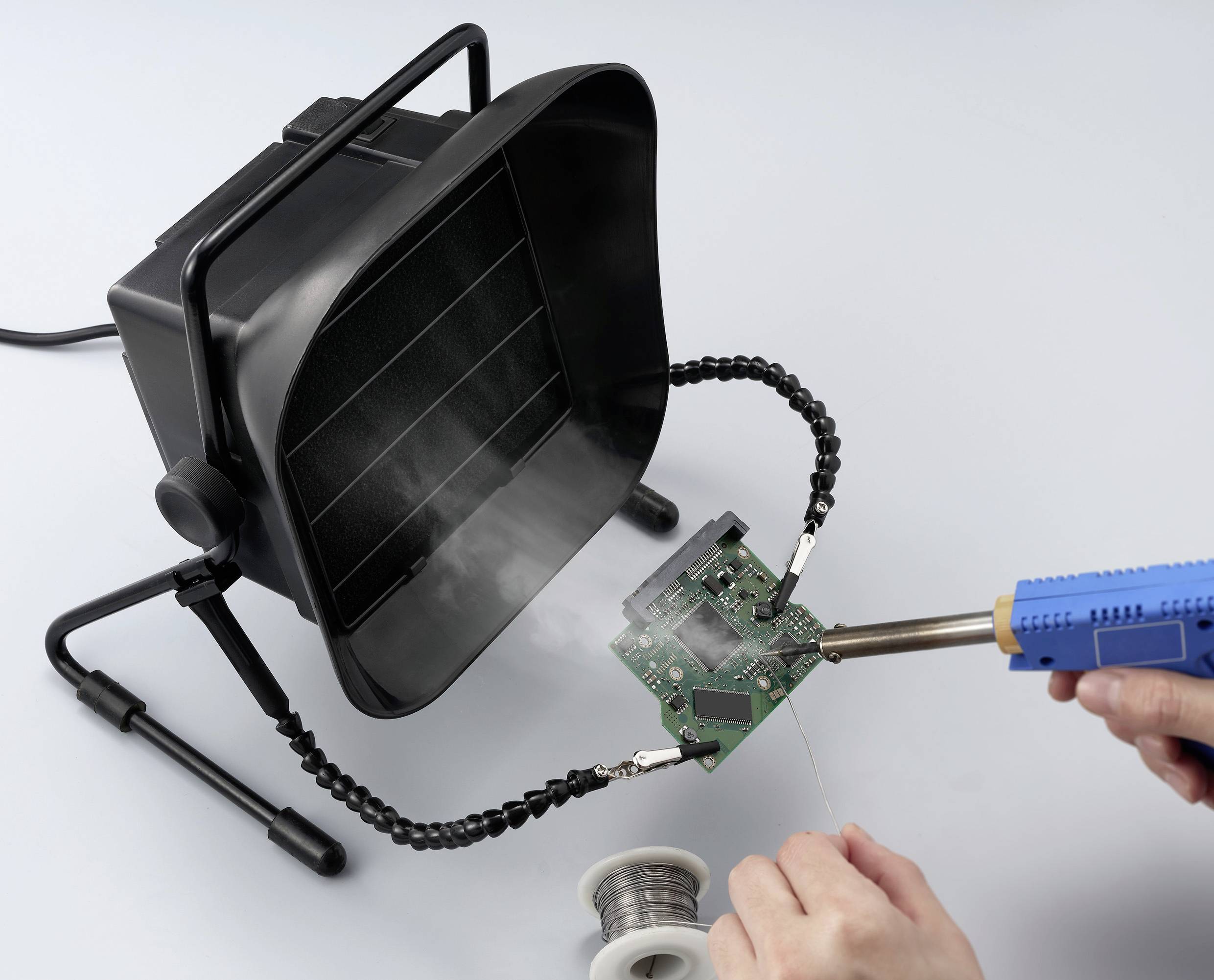 Eine Person lötet eine Leiterplatte mit einem Lötkolben, während ein Rauchabsauger Rauch entfernt. Im Hintergrund ist eine Rolle Lötzinn zu sehen.