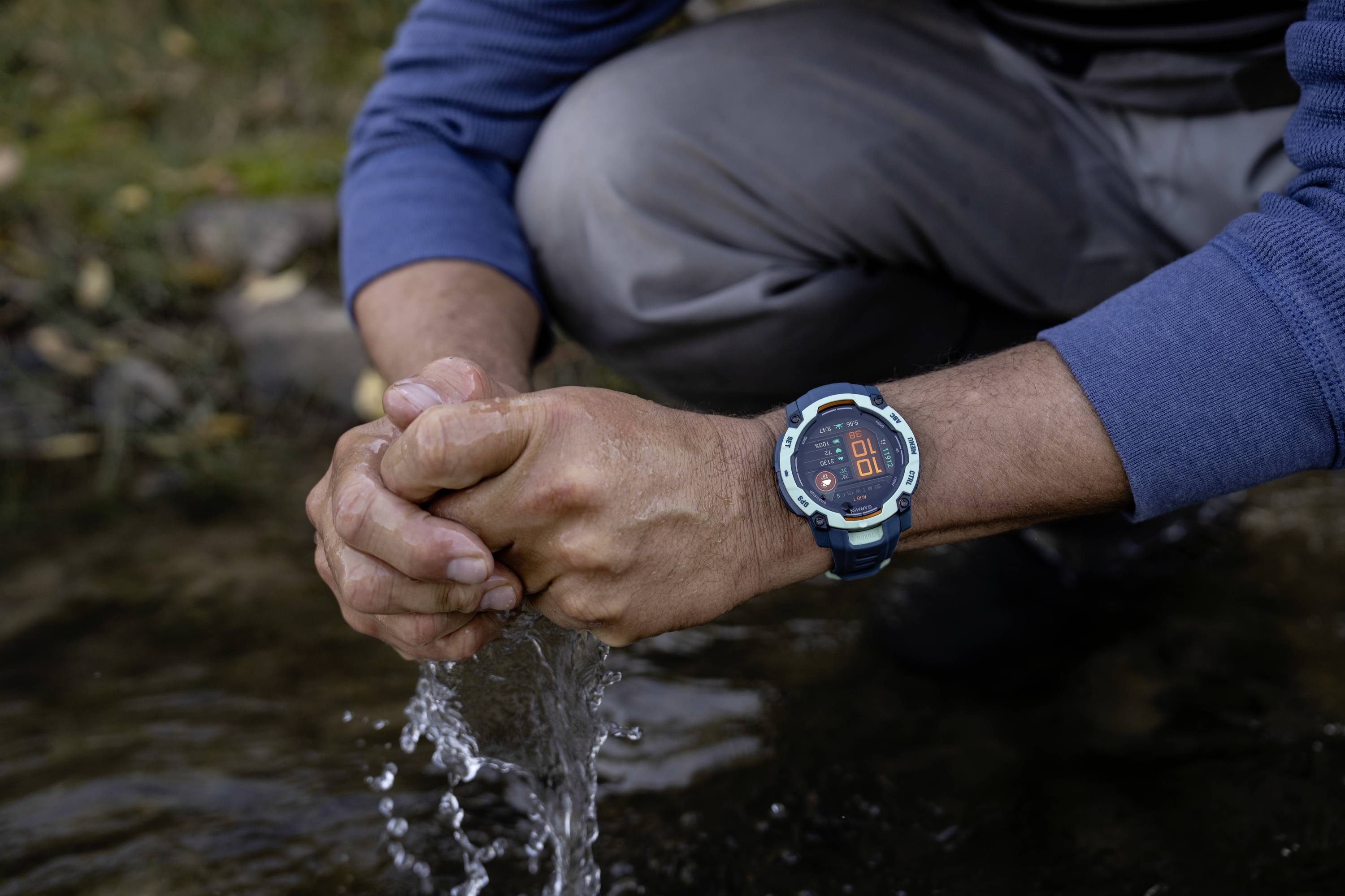 Eine Person kniet an einem Fluss und sammelt Wasser mit den Händen. Sie trägt eine Armbanduhr mit rotem Display.