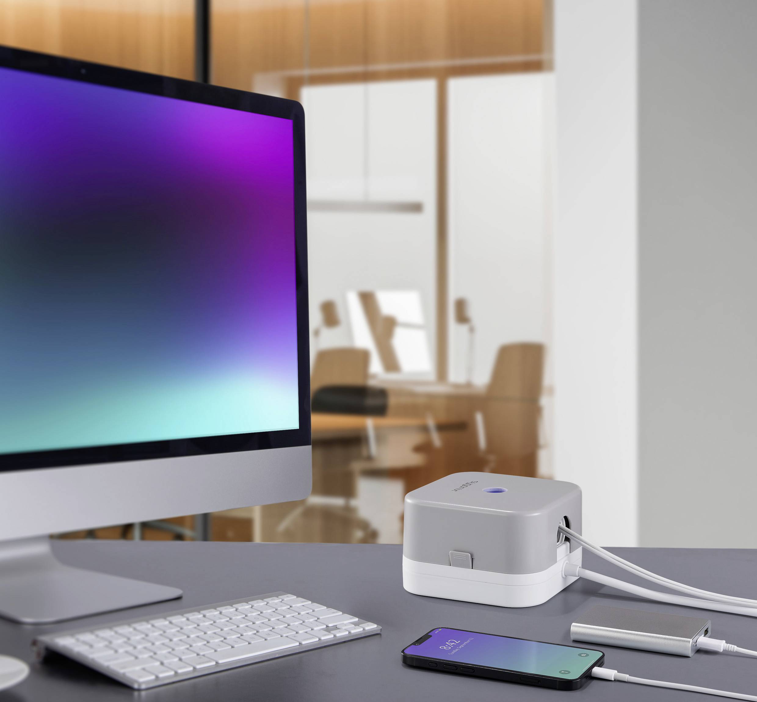 Ein Monitor, ein Smartphone und ein Würfelgerät auf einem Schreibtisch in einem modernen Büro. Das Gerät scheint als Ladegerät zu dienen.