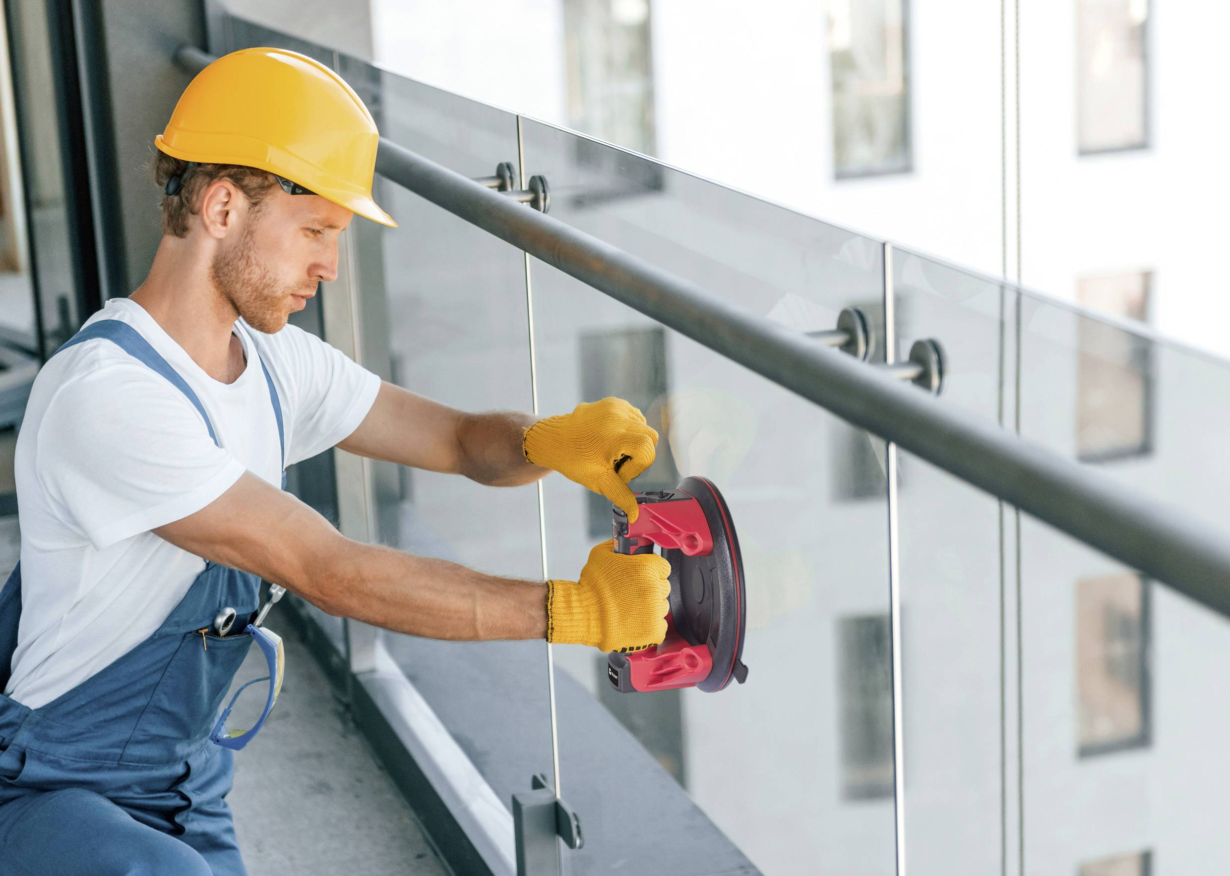 Arbeiter mit gelbem Schutzhelm montiert Glasgeländer auf einem Balkon. Er trägt Handschuhe und nutzt einen Werkzeughalter.