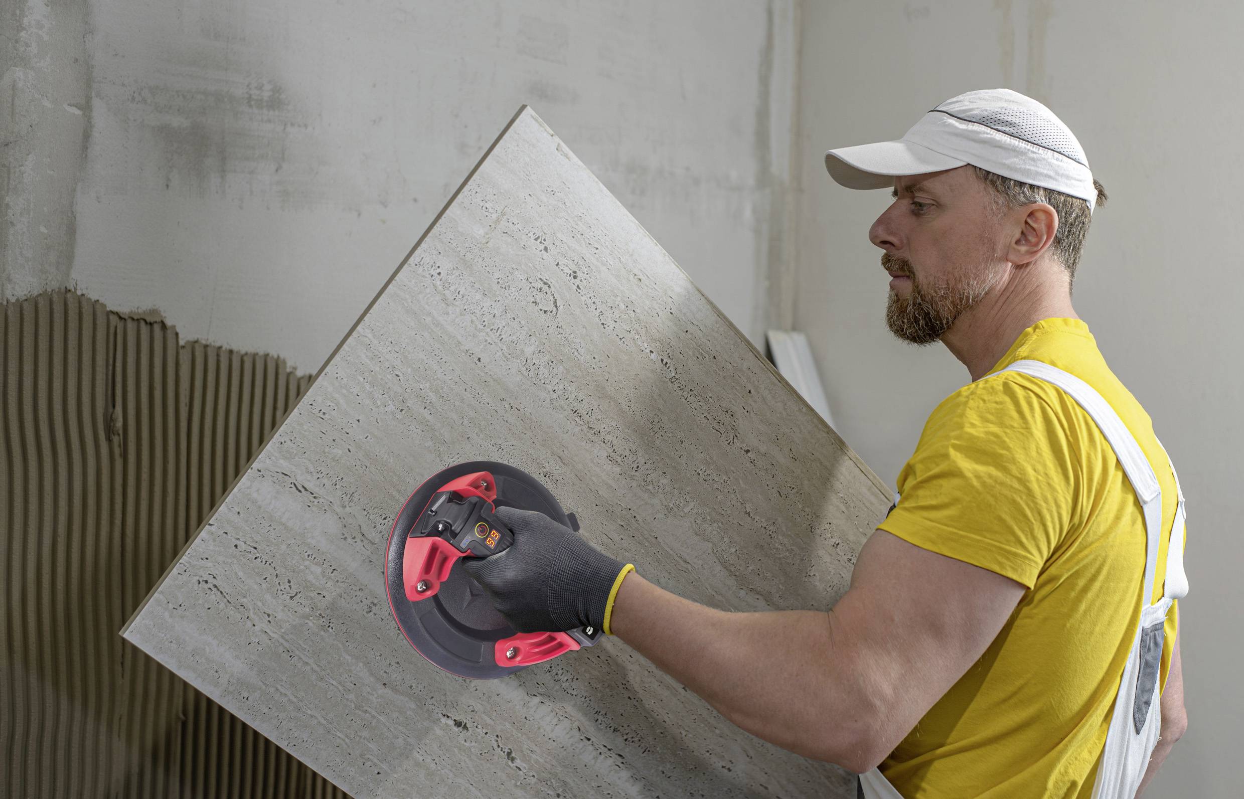 Ein Mann in gelbem T-Shirt und weißer Kappe setzt eine große Fliese an eine Wand an.