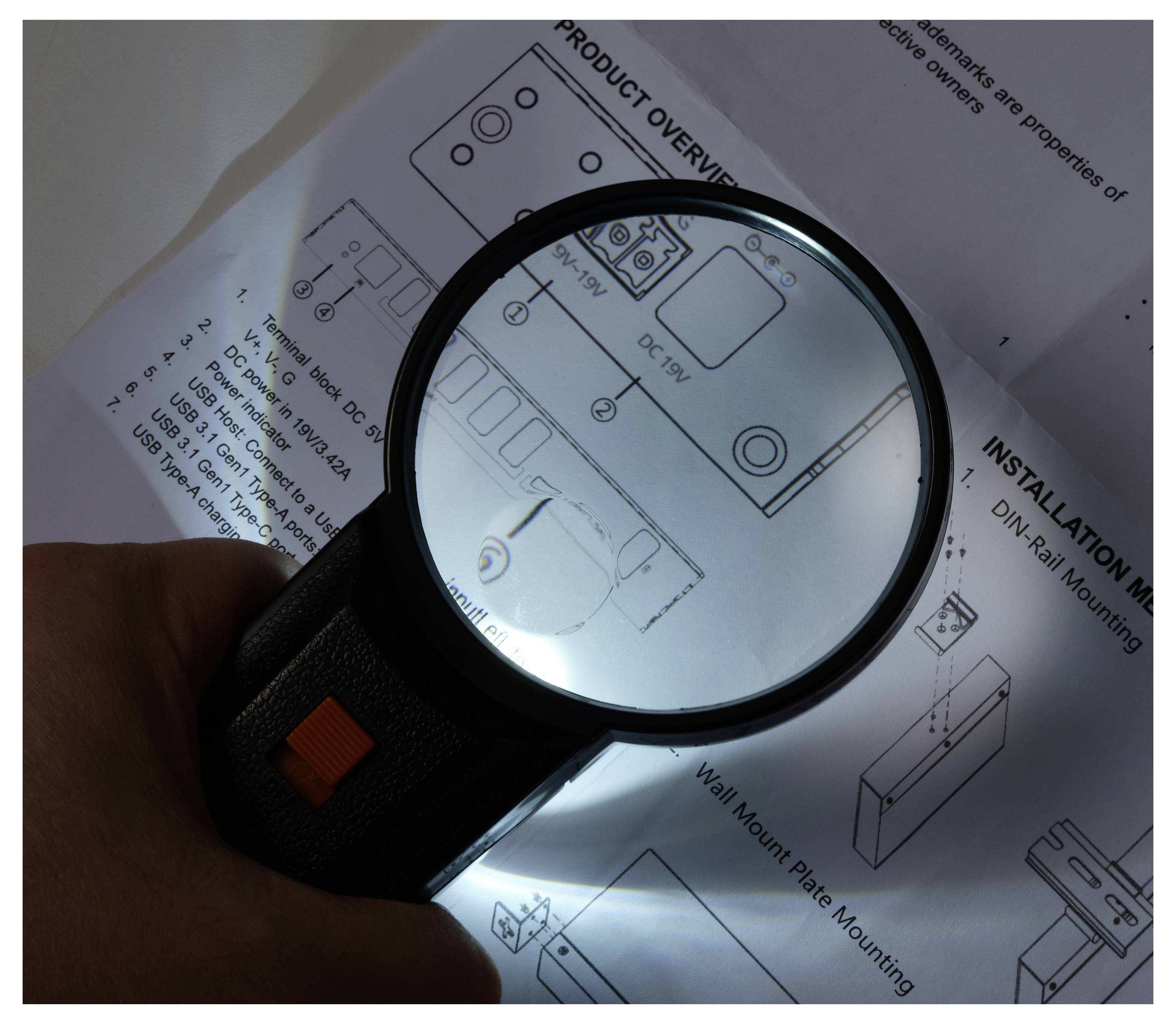 Hand holding a magnifying glass over a product instruction sheet, enlarging small assembly diagrams and text.---Hand hält eine Lupe über ein Produktinstruktionsblatt und vergrößert kleine Montage-Diagramme und Text.