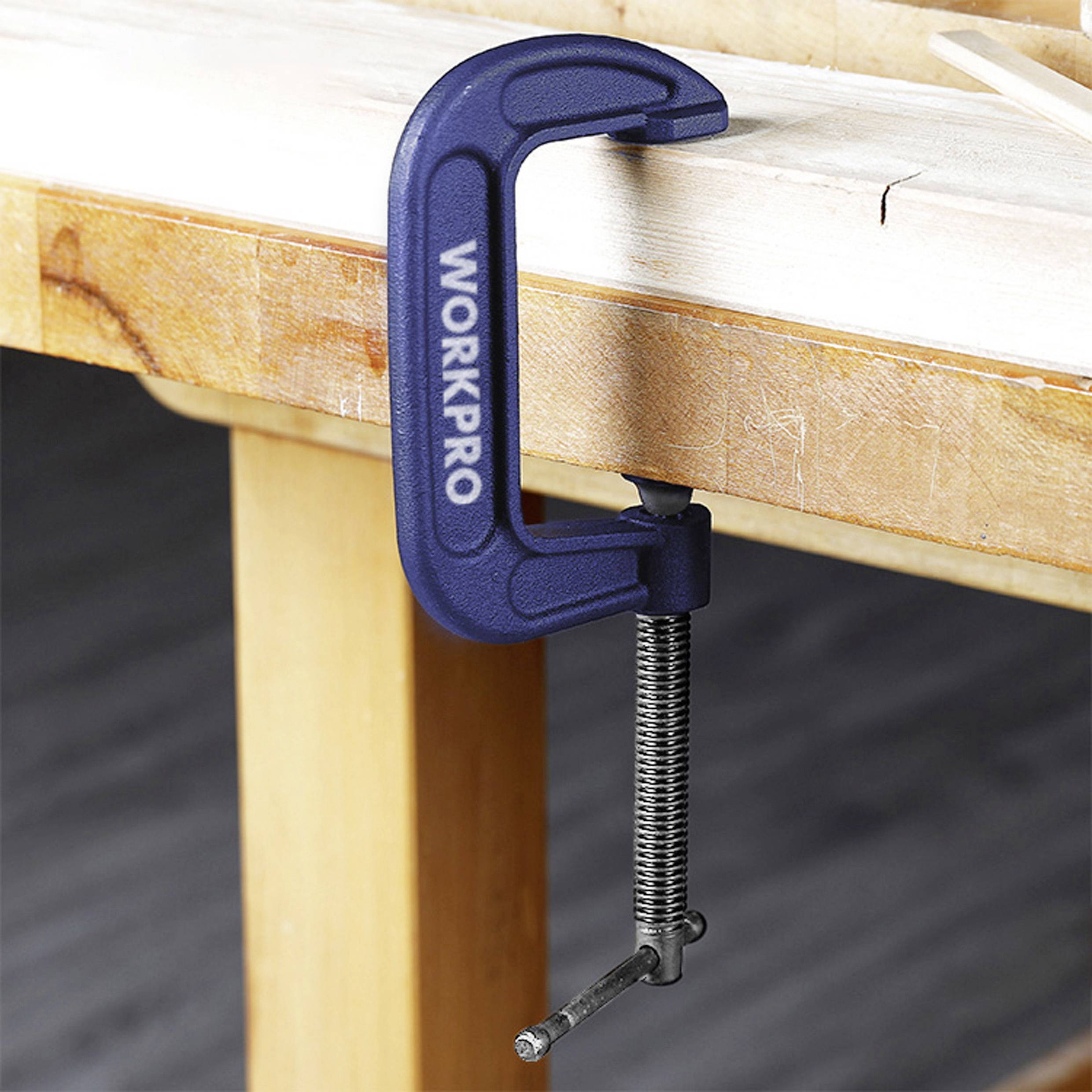 Eine blaue Schraubzwinge hält ein Stück Holz an einer Werkbank fest. Die Zwinge trägt den Aufdruck 'WORKPRO'.
