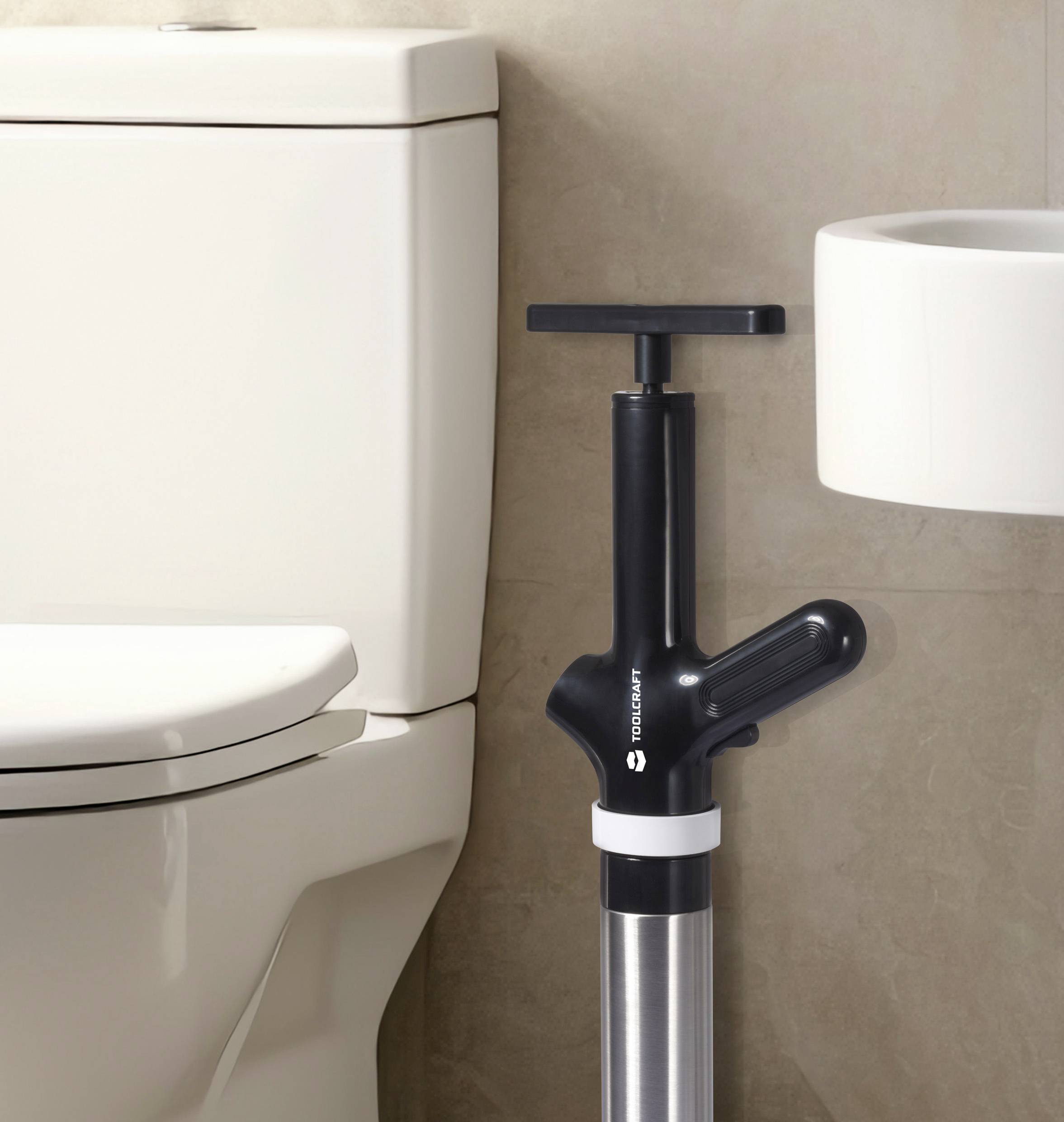 Eine Toilettenpumpe steht neben einer Toilette, bereit zur Nutzung. Im Hintergrund ein Waschbecken.