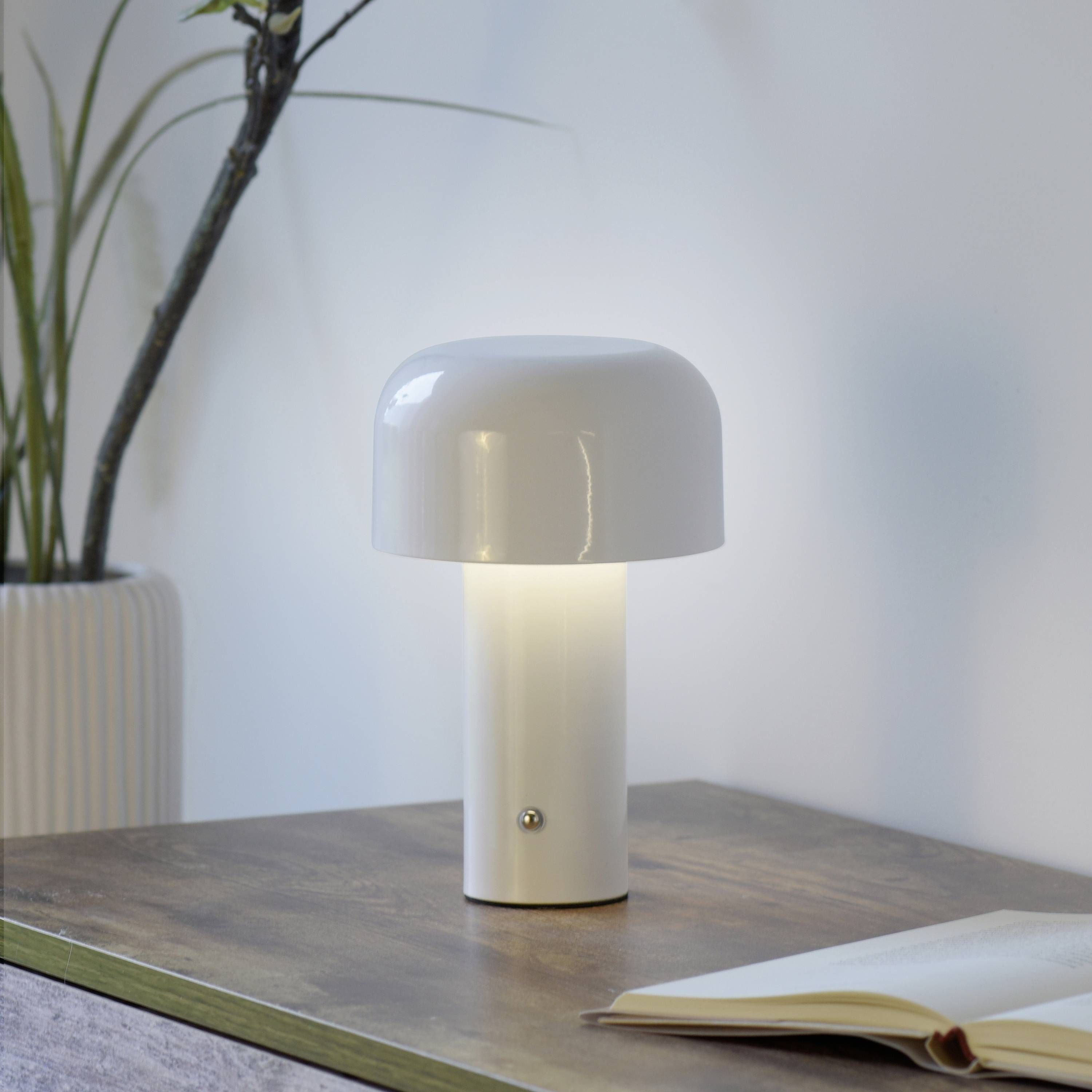 Eine weiße Tischlampe leuchtet auf einem Holztisch neben einem geöffneten Buch. Im Hintergrund steht eine Pflanze in einem Topf.