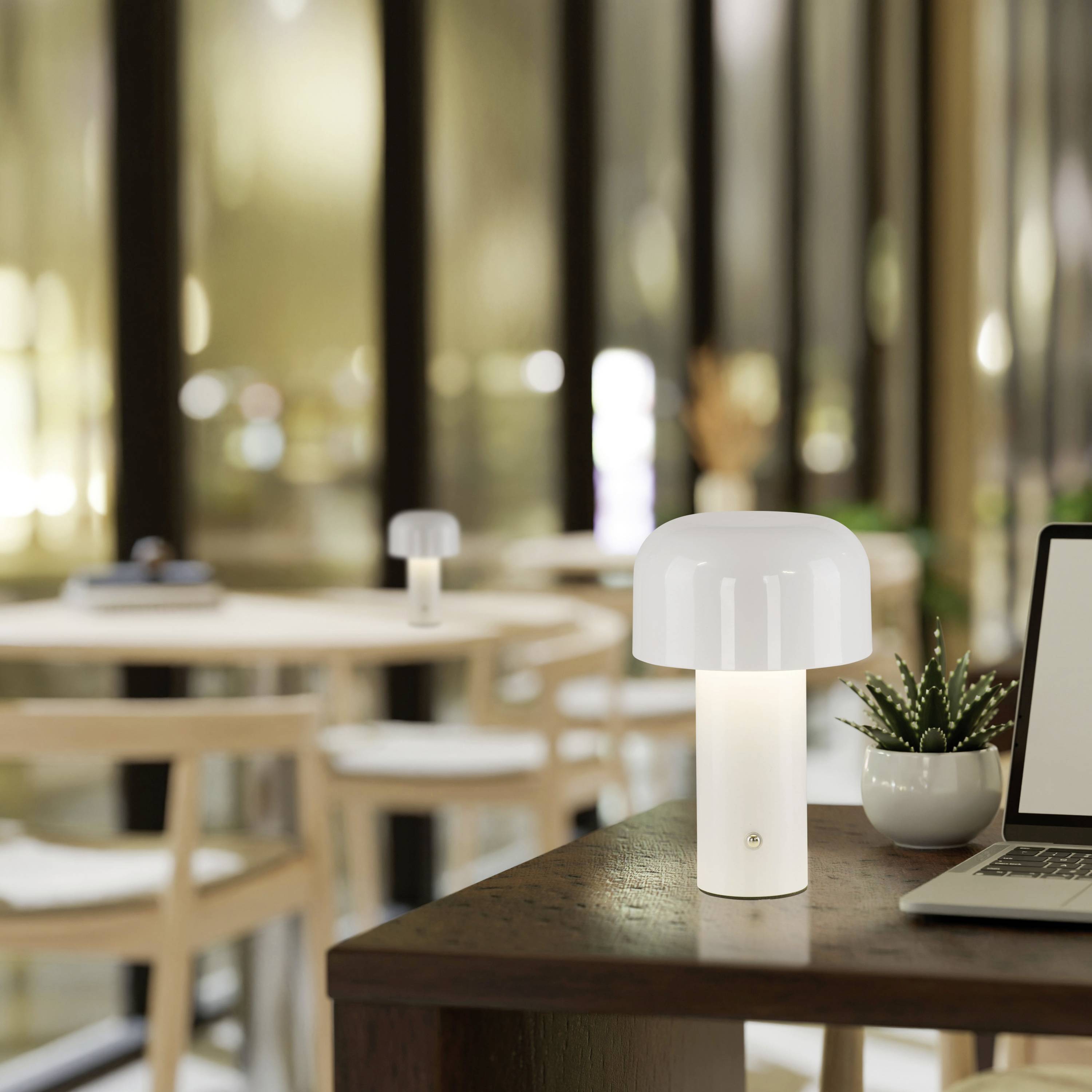 Eine kleine, weiße Tischlampe erhellt einen Holztisch in einem modern eingerichteten Café. Im Hintergrund sind unscharfe Tische und Stühle sichtbar.