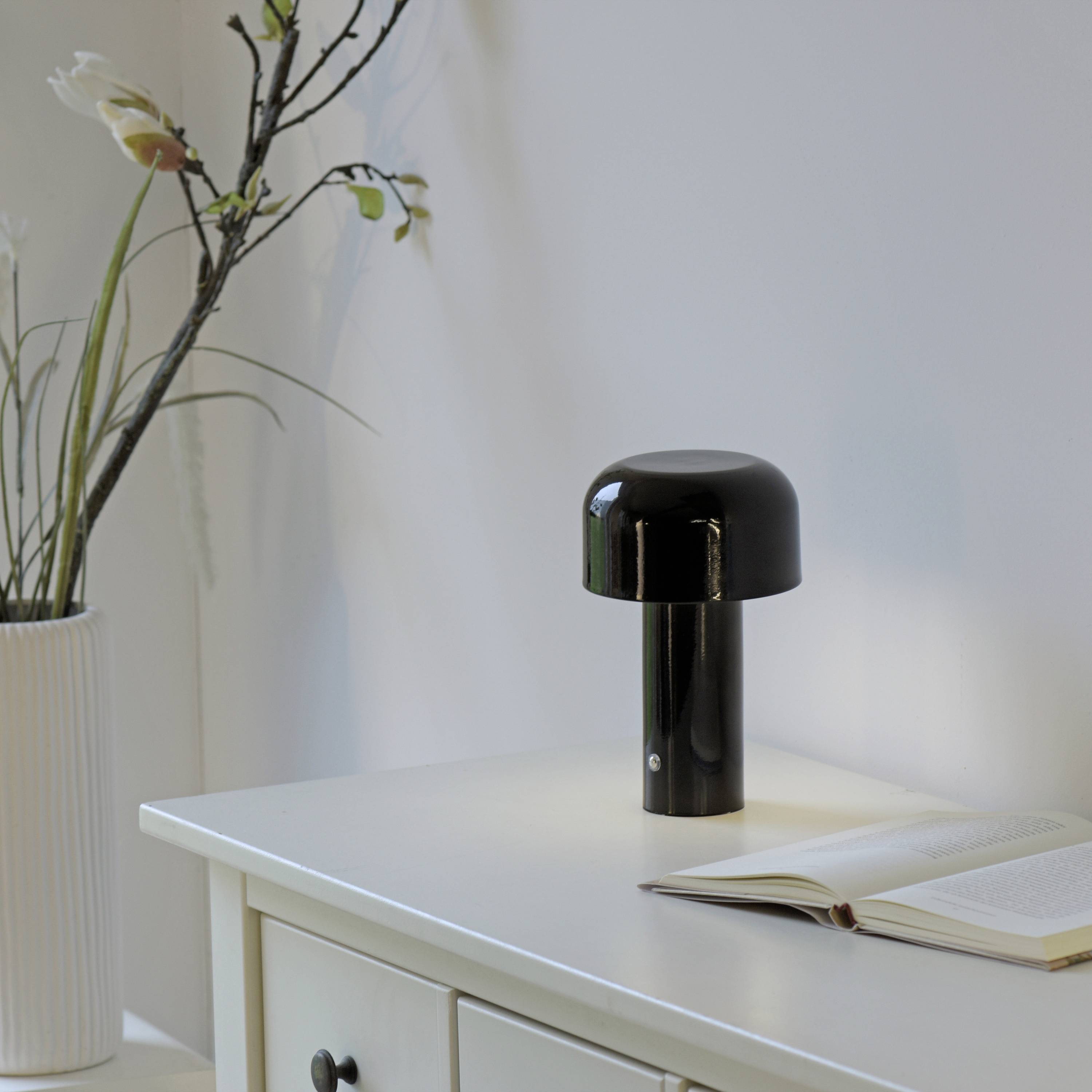 Schwarze Lampe auf weißem Tisch neben einem aufgeschlagenen Buch und einer Vase mit Zweigen, in einem ruhigen Raum.