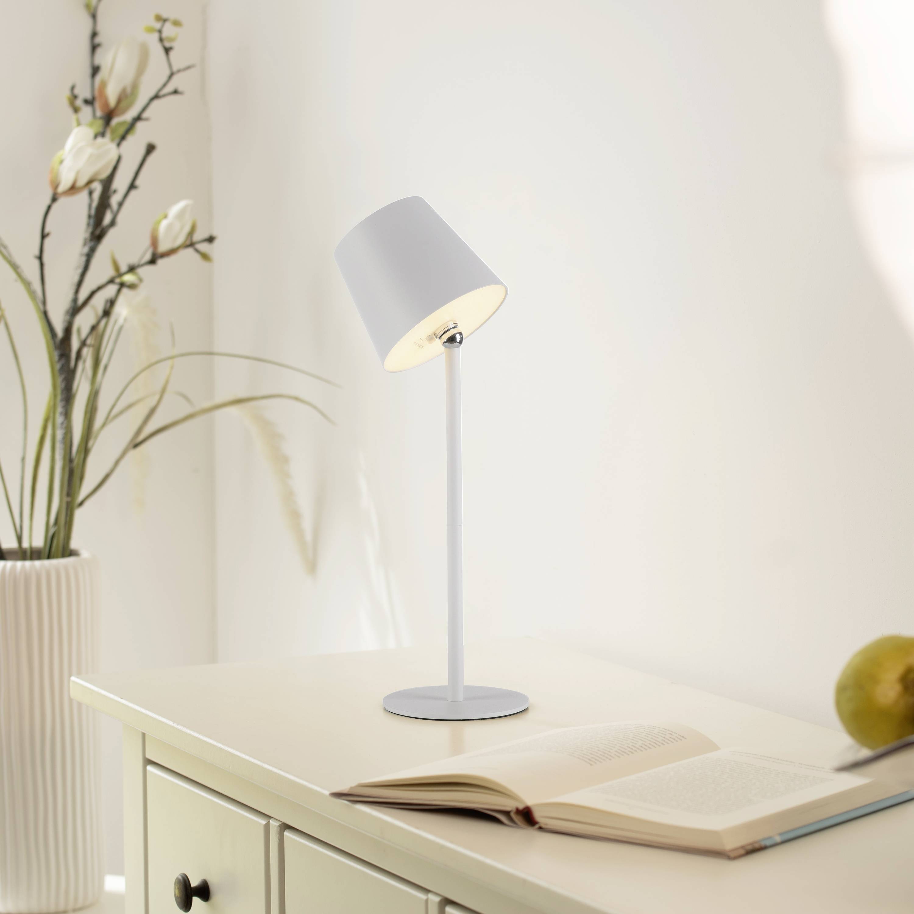 Eine geöffnete Buchseite auf einem Tisch neben einer eingeschalteten Tischlampe. Daneben steht eine Vase mit Zweigen.