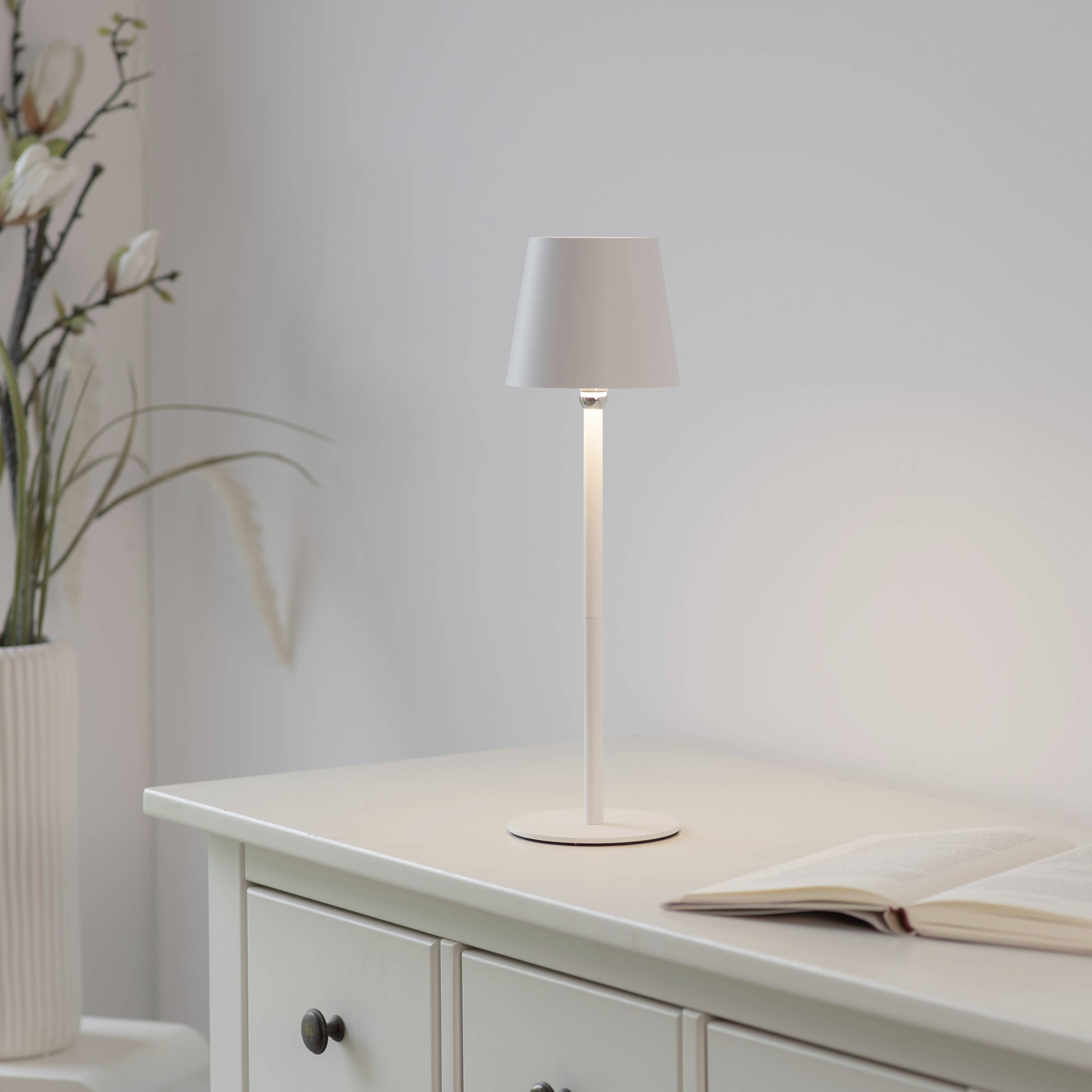 Eine minimalistische Tischlampe beleuchtet ein aufgeschlagenes Buch auf einem weißen Sideboard. Links steht eine Vase mit Blumen.