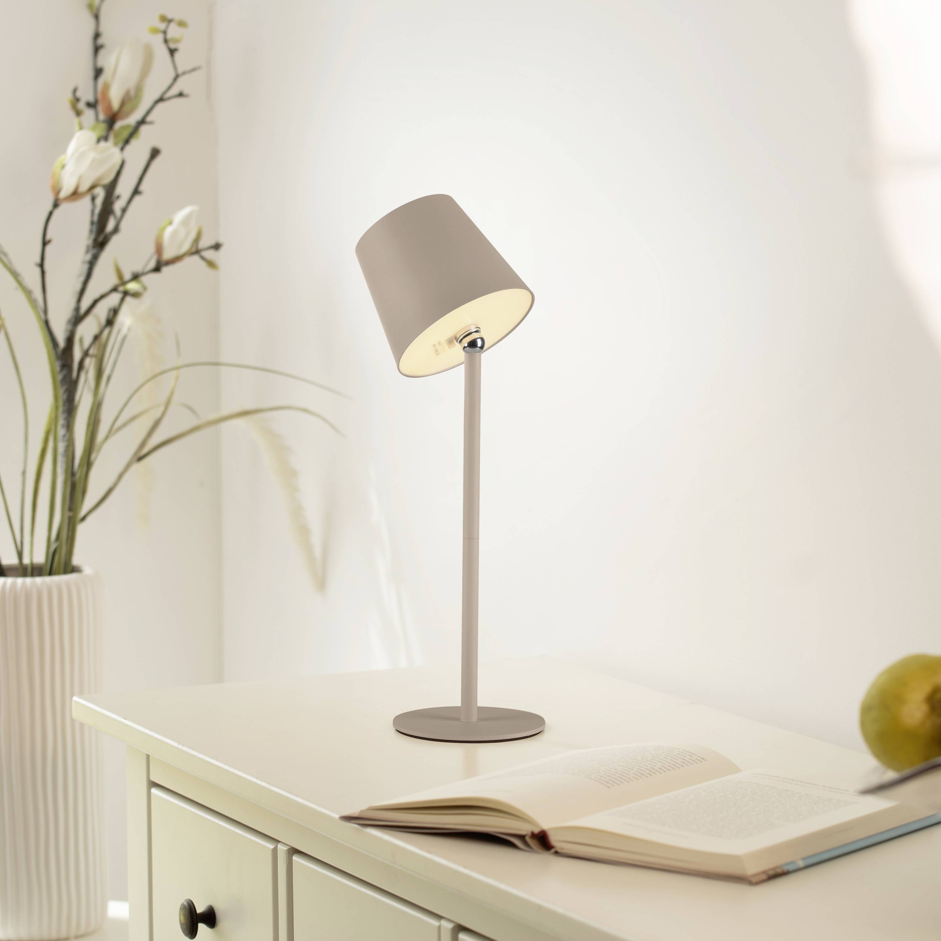 Eine Tischlampe mit warmem Licht beleuchtet ein offenes Buch auf einem Tisch. Im Hintergrund steht eine Vase mit weißen Blumen.