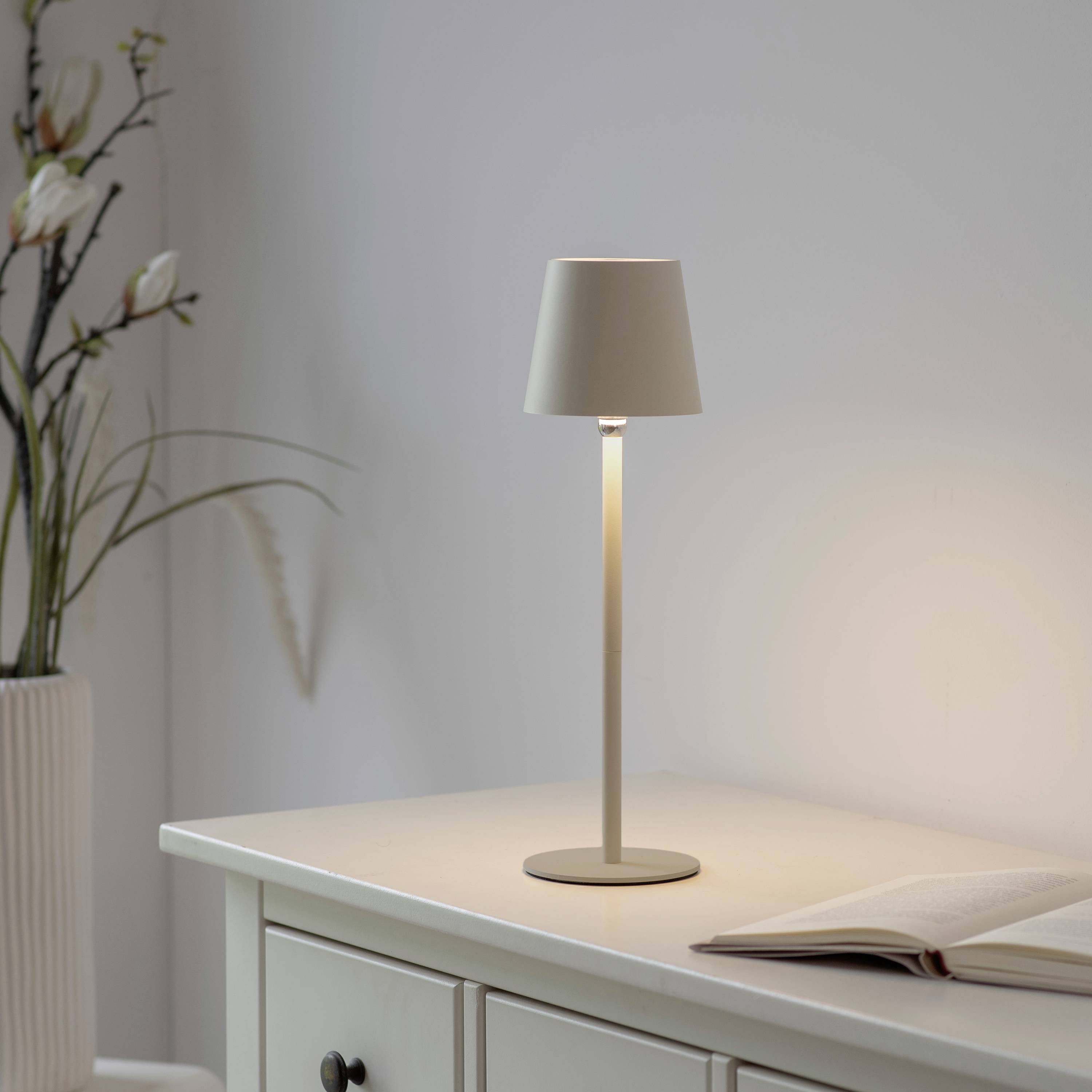 Lampe auf Tisch neben offenem Buch und Vase mit Zweigen, schafft eine ruhige und einladende Atmosphäre.