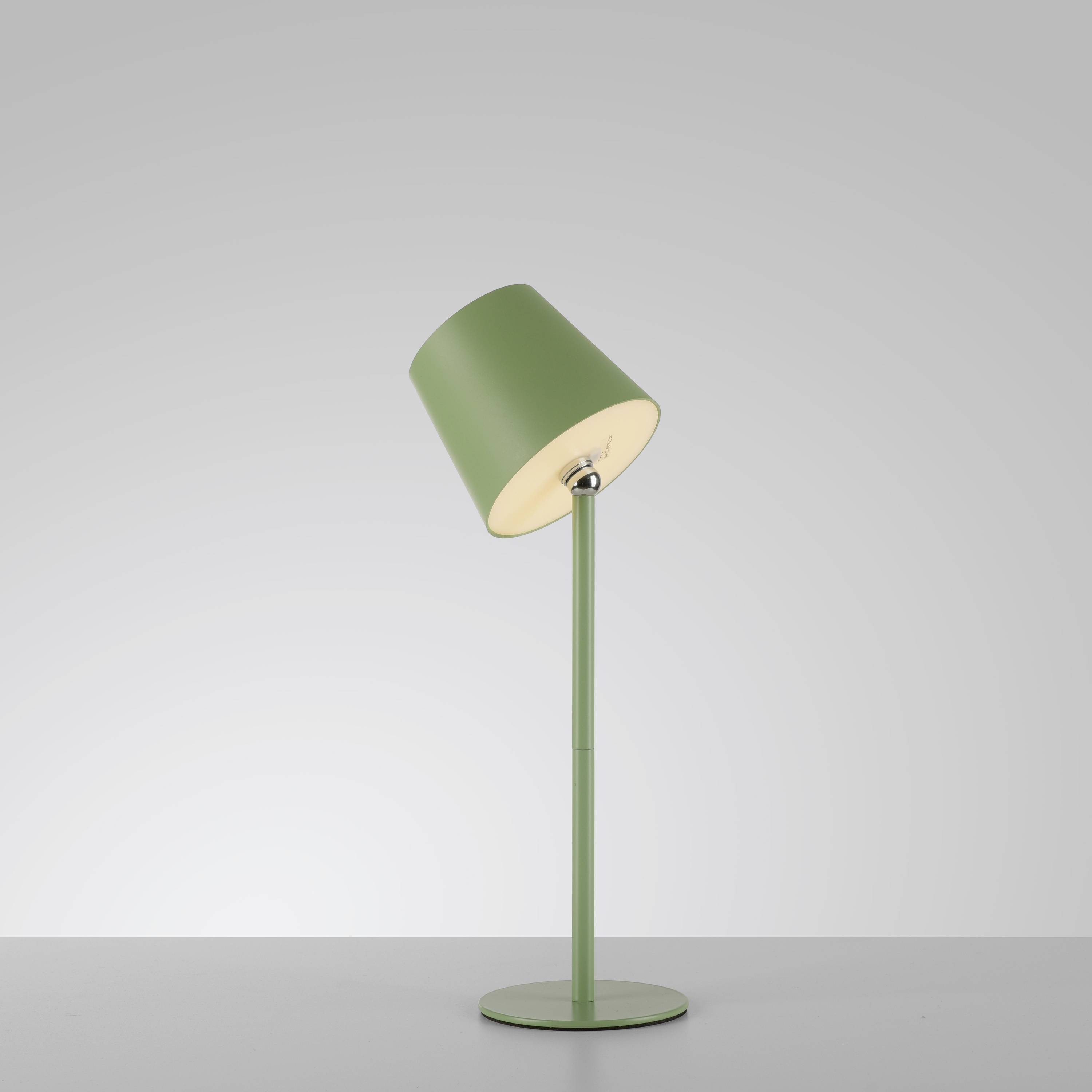 Eine grüne Tischlampe mit schrägem Schirm steht auf einem grauen Hintergrund. Sie ist eingeschaltet und leuchtet hell.