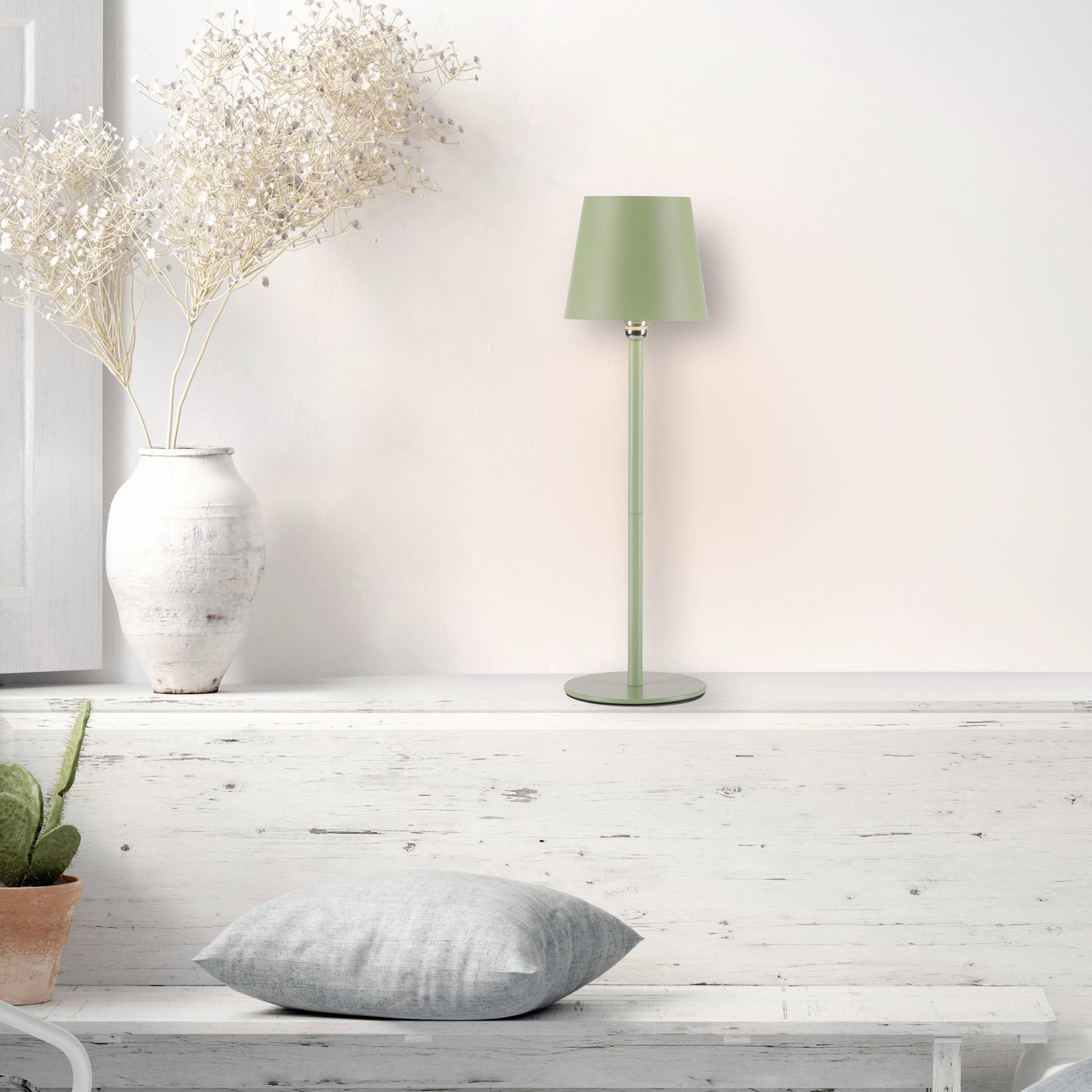 Eine grüne Tischlampe steht auf einem weißen Holzregal neben einer weißen Vase mit getrockneten Blumen und einem grauen Kissen.