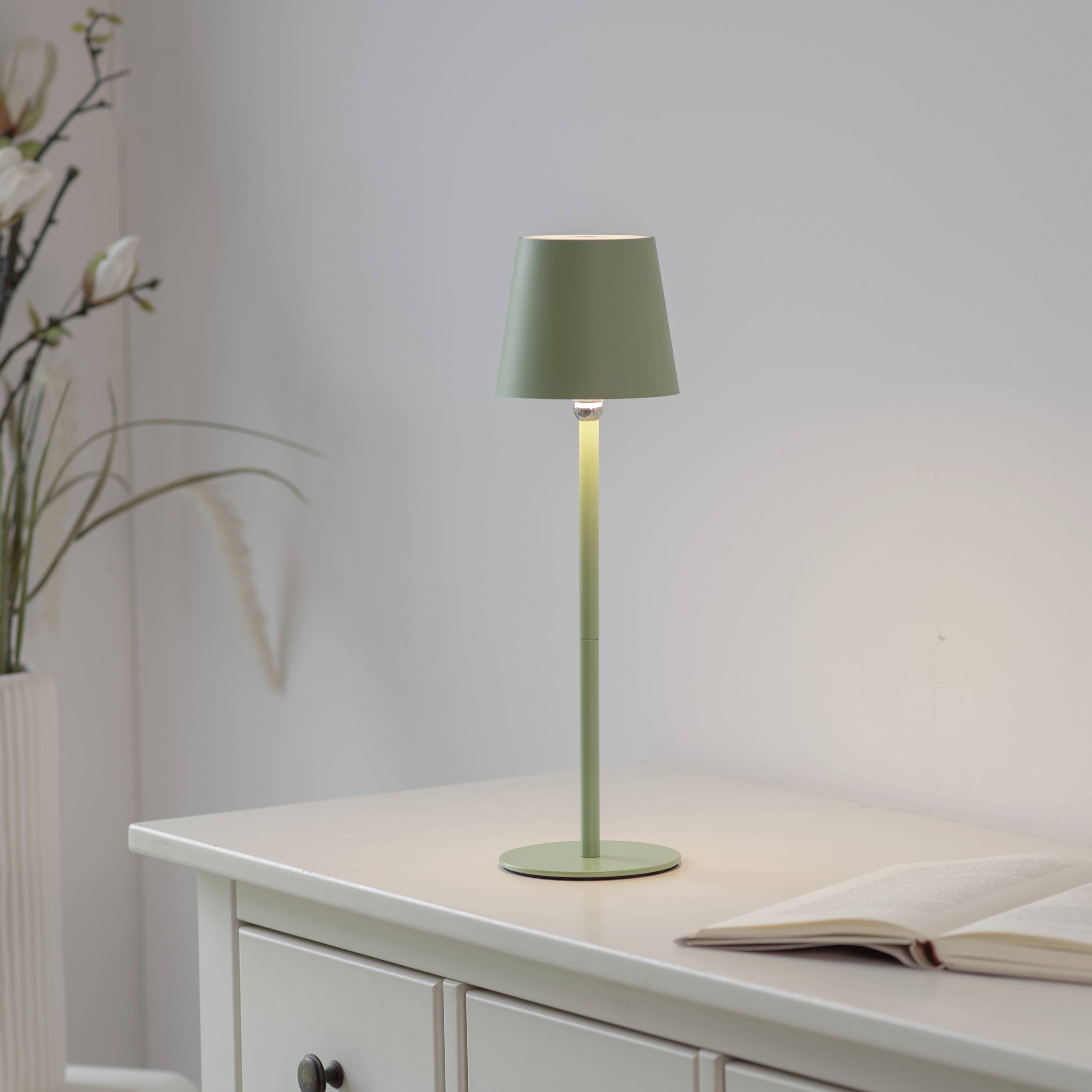 Eine grüne Tischlampe auf einem weißen Tisch beleuchtet ein geöffnetes Buch, das im Hintergrund steht, mit Blumen daneben.