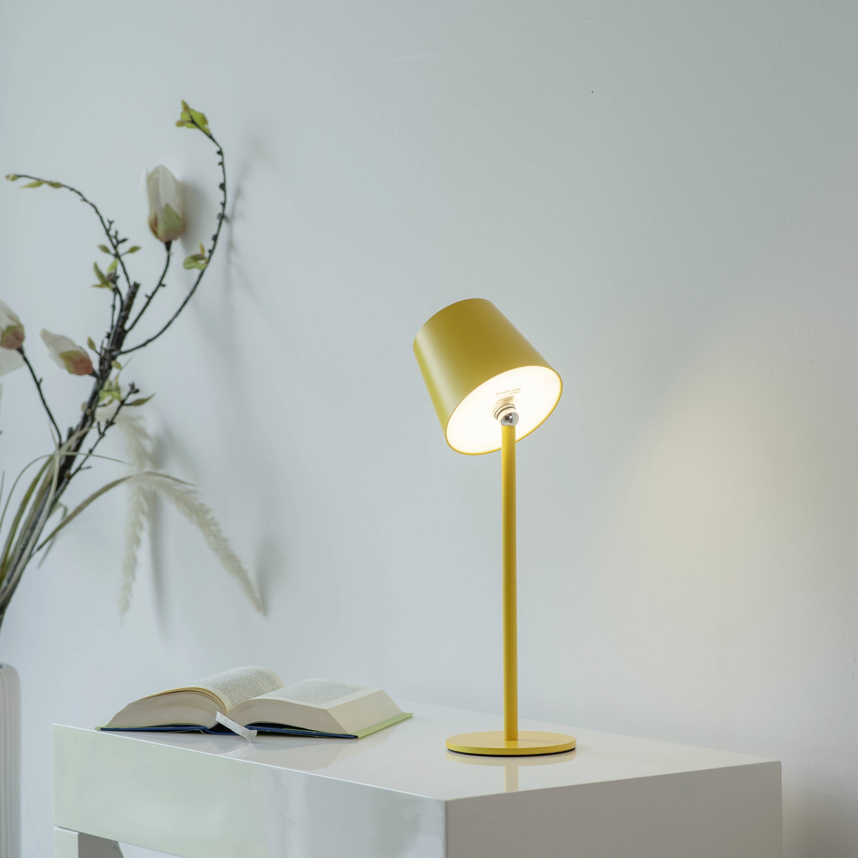 Moderne gelbe Tischlampe auf weißem Tisch neben aufgeschlagenem Buch, im Hintergrund blühende Blumen an der Wand.