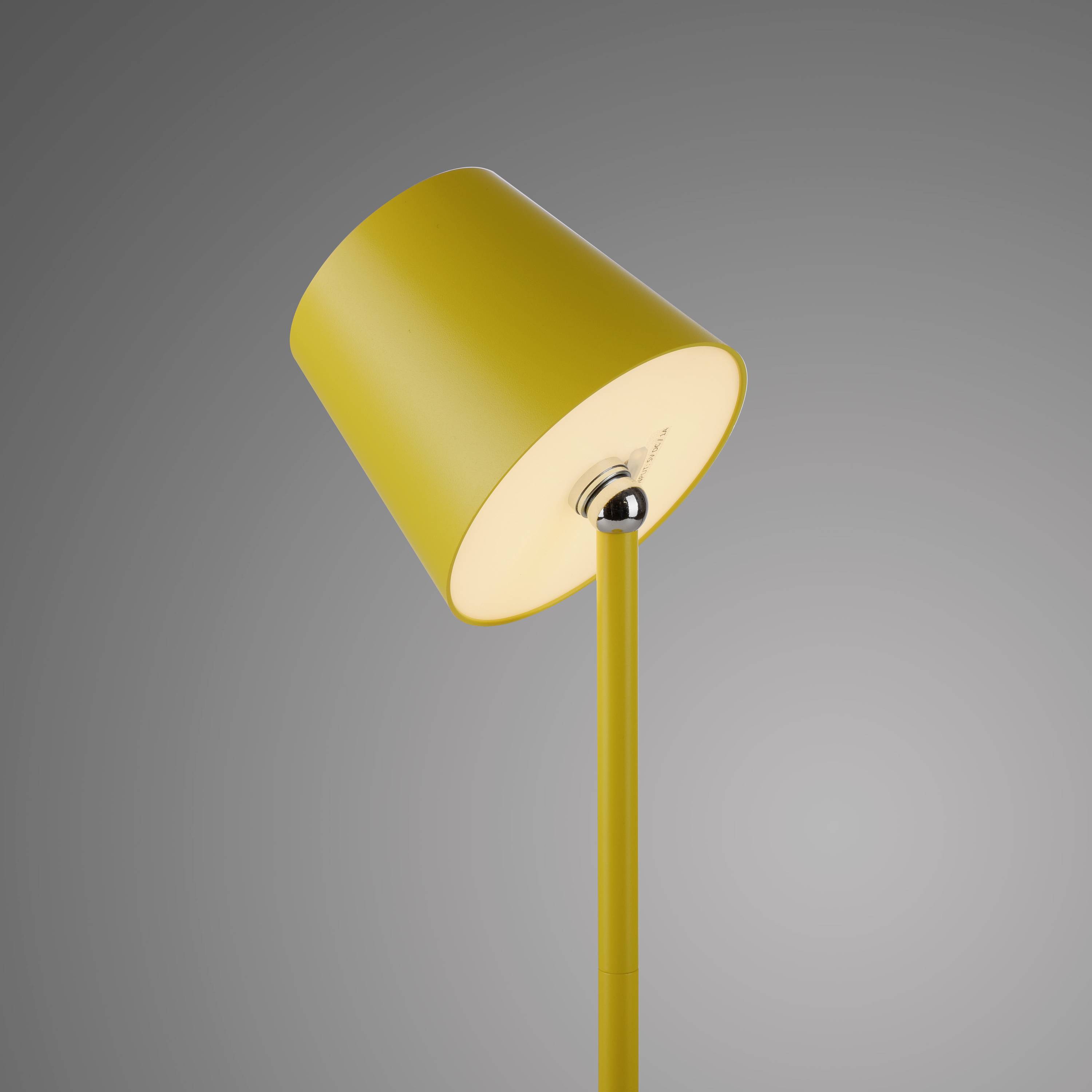 Gelbe Tischlampe mit schlichtem, modernen Design vor grauem Hintergrund. Der Lampenschirm ist zylinderförmig und leicht geneigt.