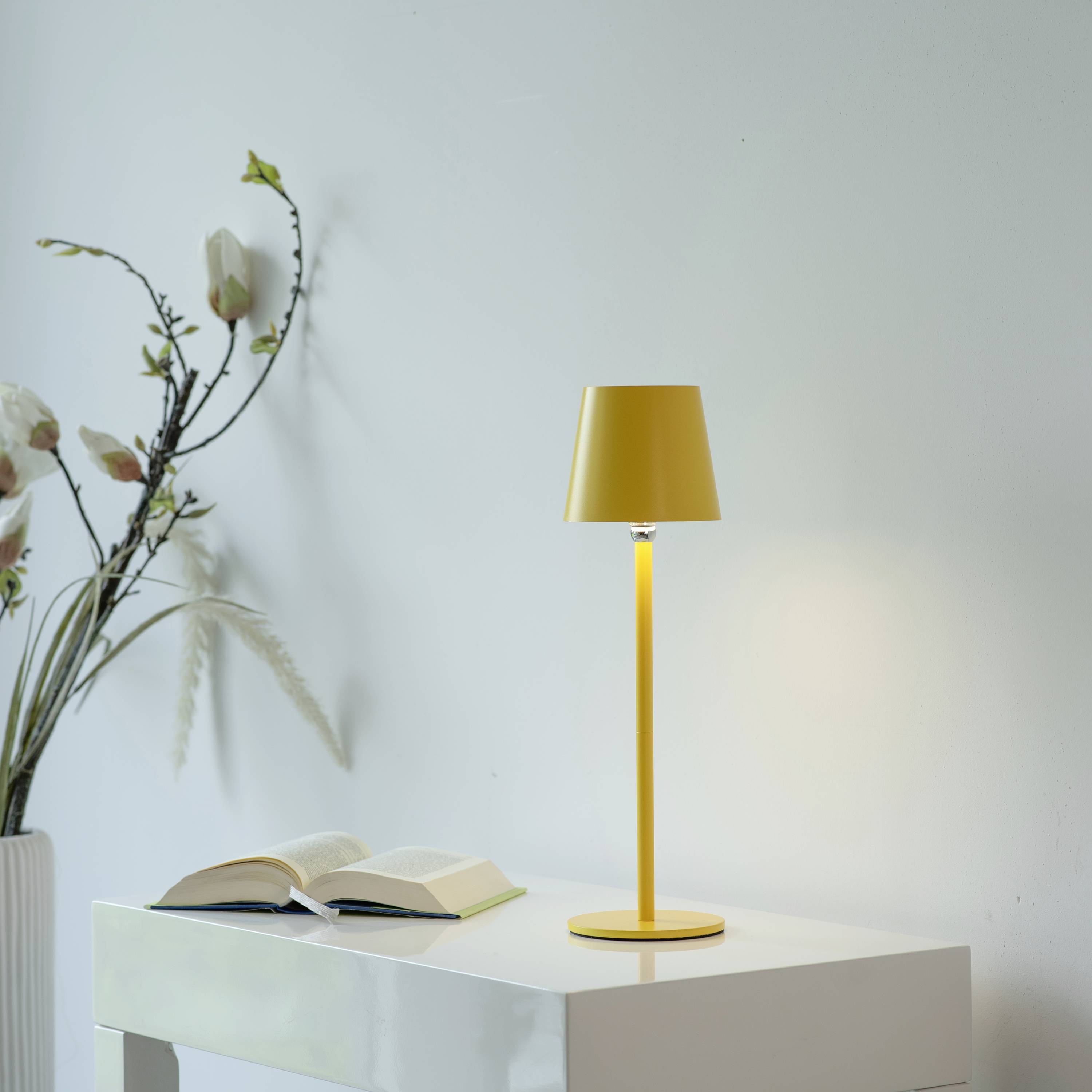 Ein gelber Tischlampe steht auf einem weißen Tisch neben einem geöffneten Buch. Im Hintergrund ist eine Vase mit Blumen zu sehen.