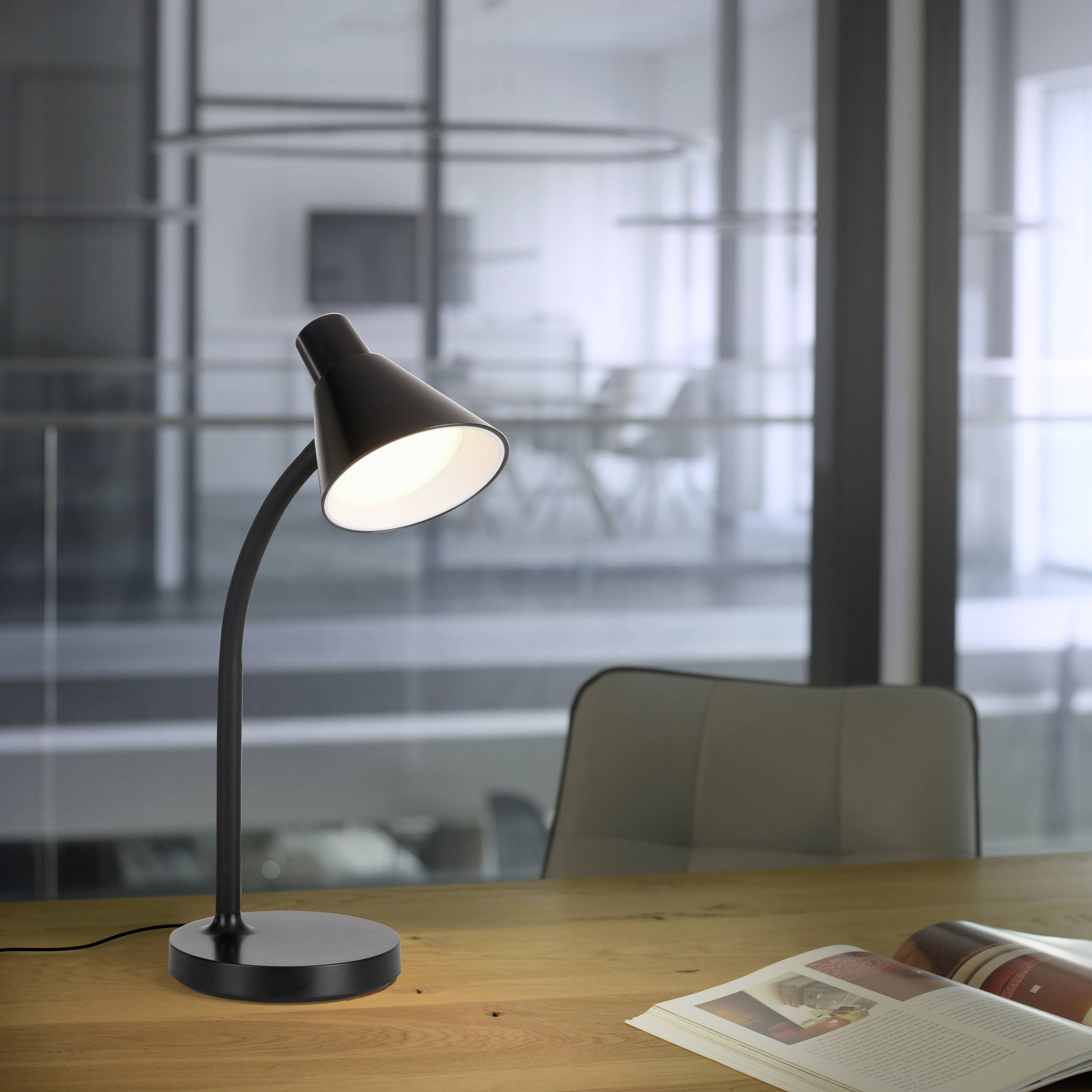 Eine schwarze Tischlampe beleuchtet einen Schreibtisch mit einem offenen Buch. Im Hintergrund sind verschwommene Büromöbel zu sehen.