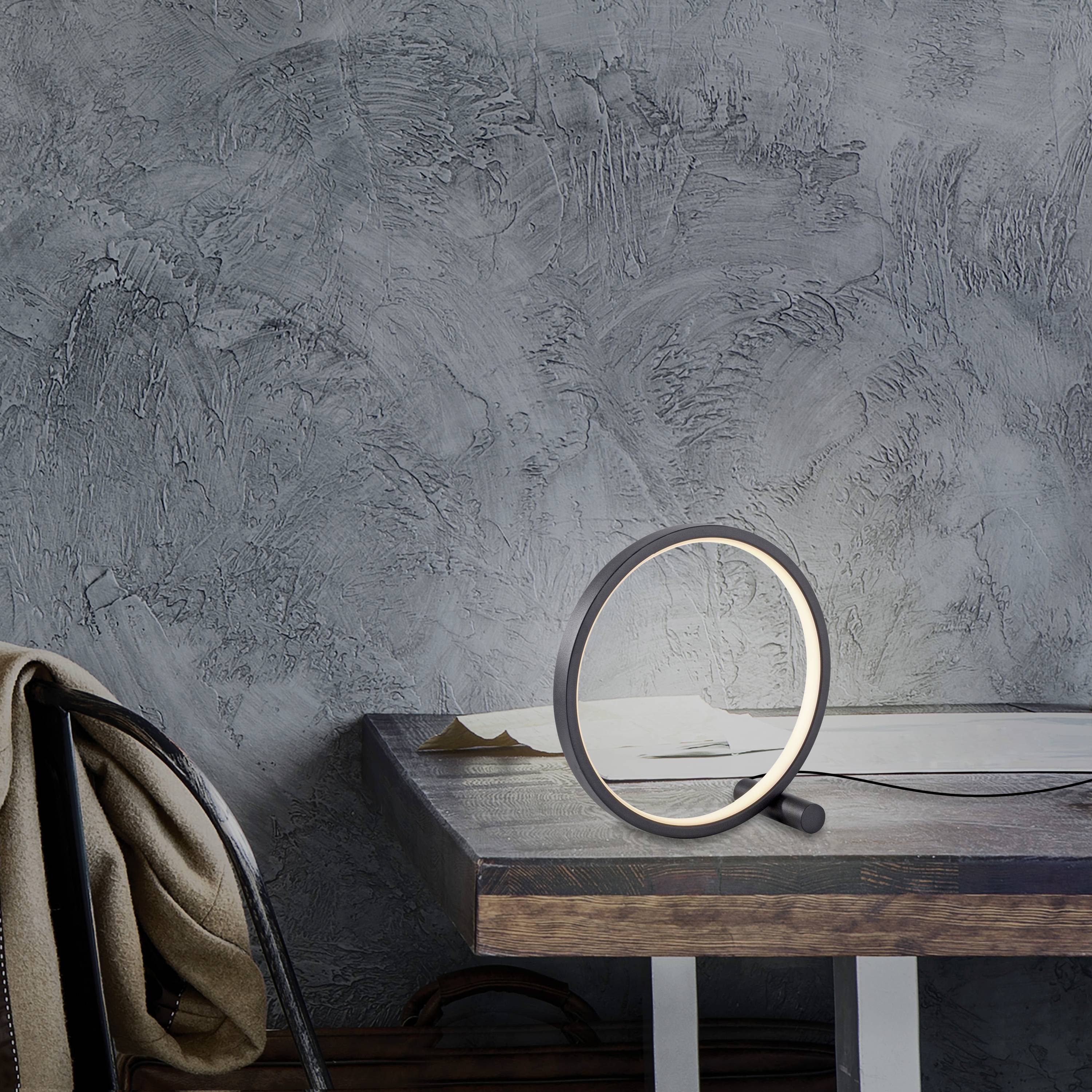 Eine runde Tischleuchte steht auf einem Holztisch vor einer strukturierten grauen Wand. Daneben liegen ein Buch und einige Papierblätter. Ein Stuhl ist teilweise sichtbar.