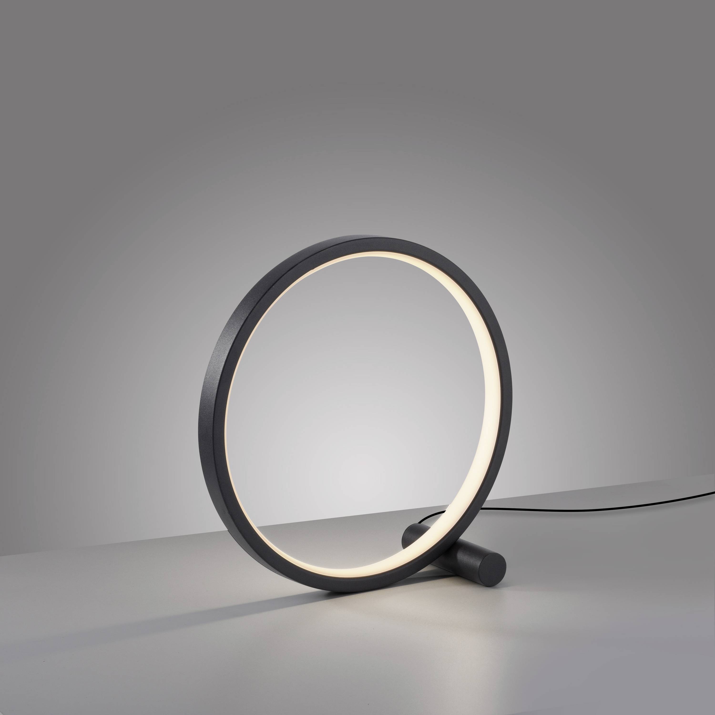 Eine moderne Tischlampe mit kreisförmigem Leuchtkörper aus schwarzem Metall, leuchtet sanft auf einer grauen Oberfläche.