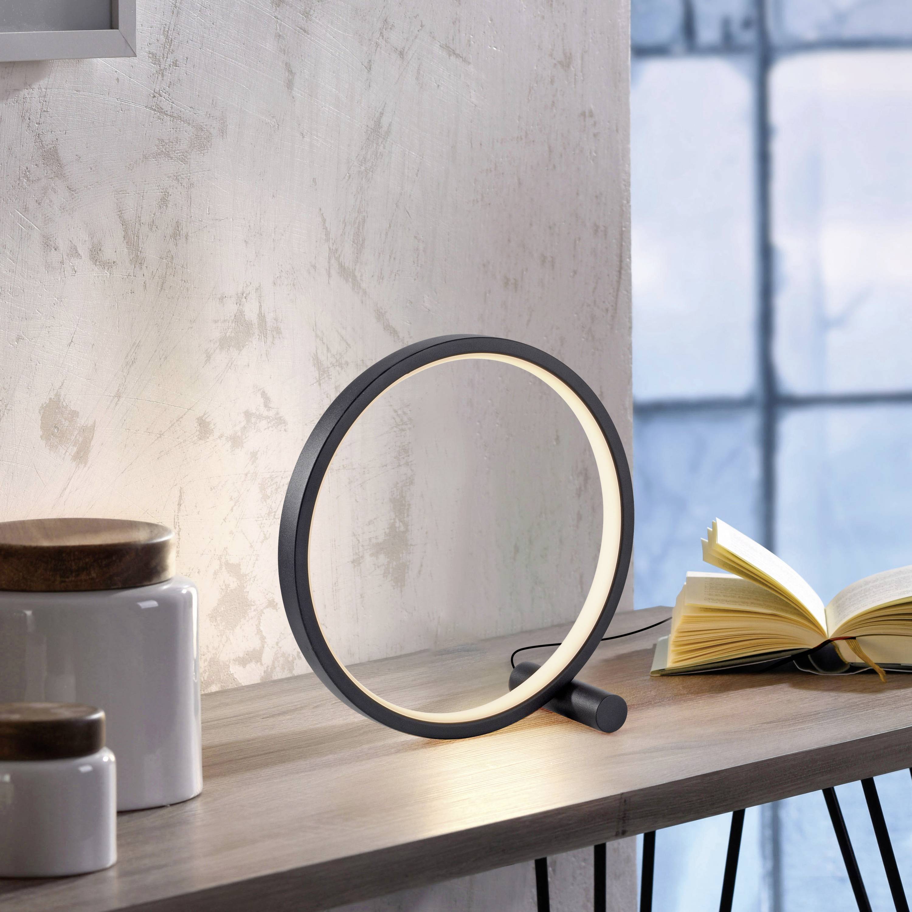 Eine runde LED-Lampe steht auf einem Regal neben gestapelten Büchern und Keramikbehältern. Im Hintergrund ist ein Fenster zu sehen.