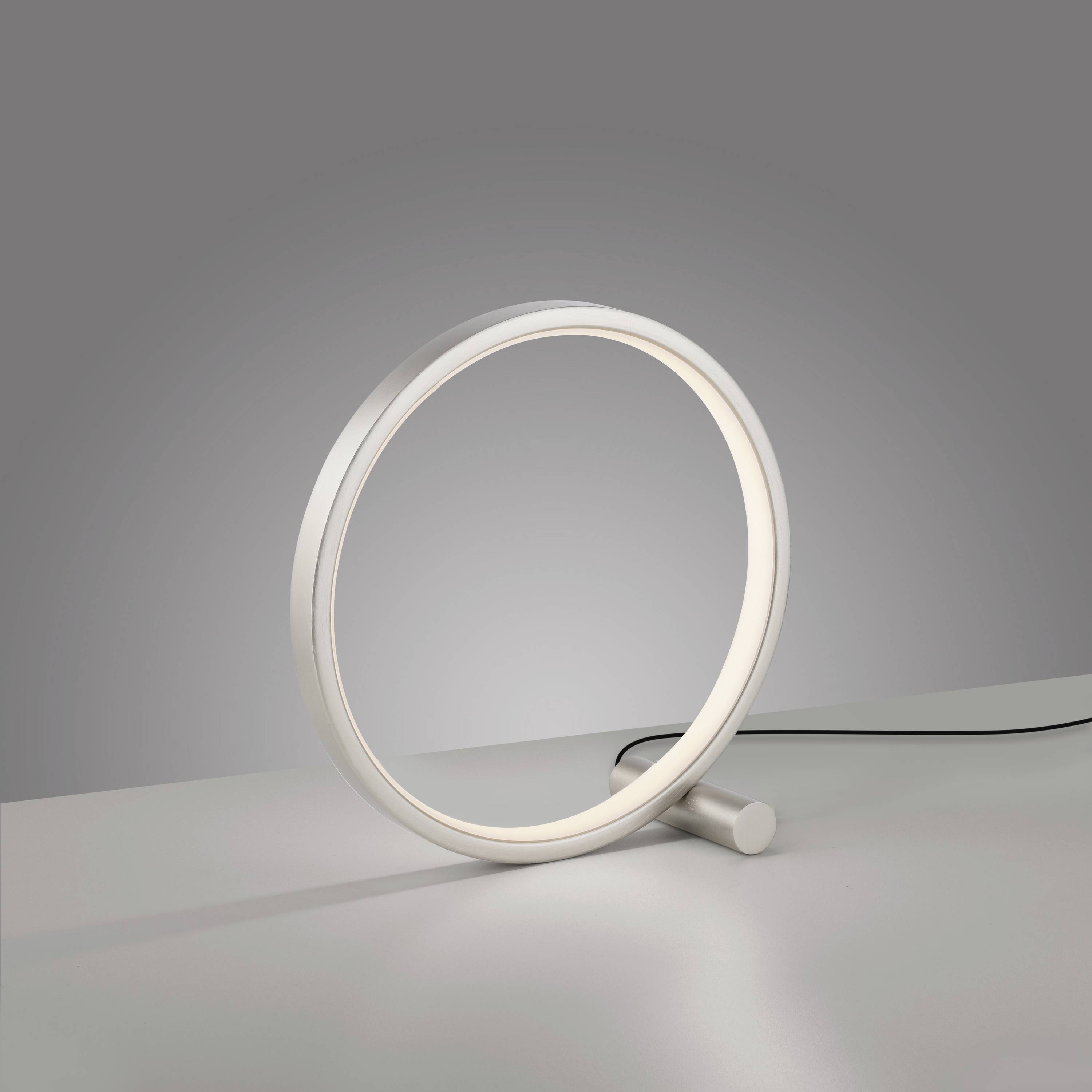 Eine moderne Tischlampe in Ringform mit warmem Licht, steht auf einem grauen Untergrund. Sie hat ein minimalistisches, schlankes Design.
