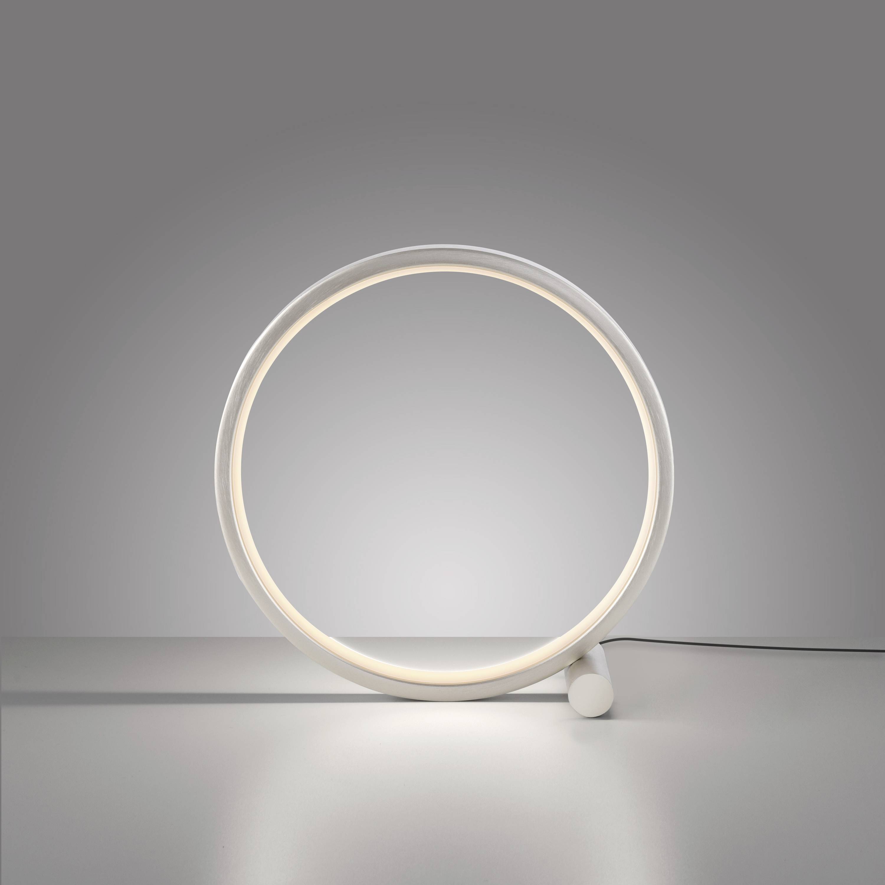 Eine moderne Lampe in Form eines leuchtenden Rings auf einem Tisch. Der Ring strahlt ein weiches, gleichmäßiges Licht aus.