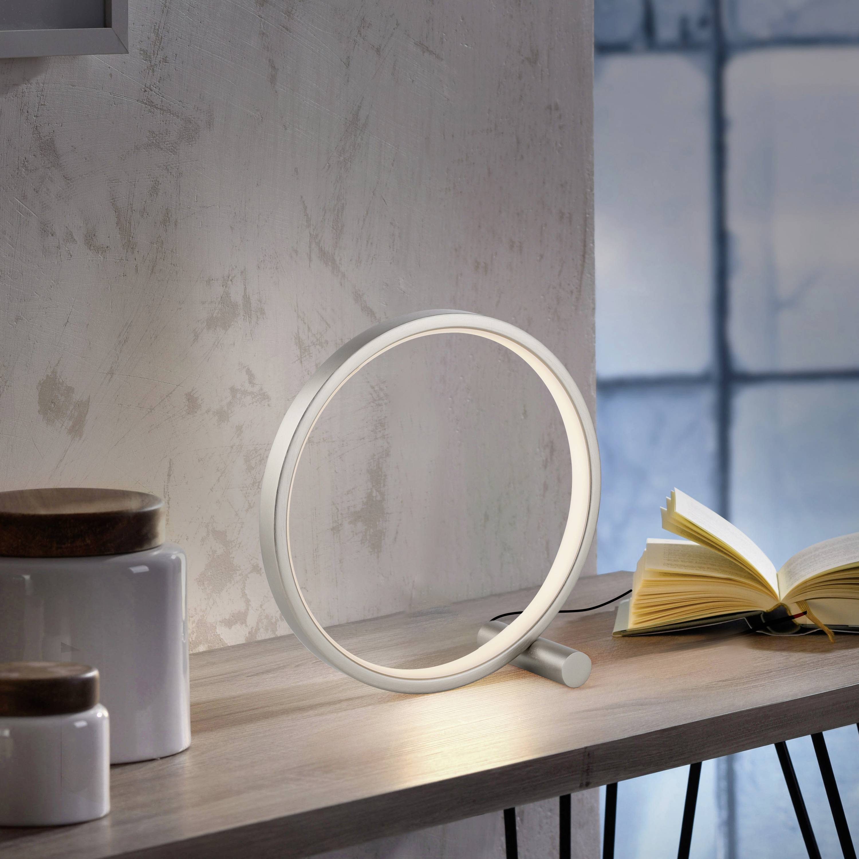 Eine runde, moderne Tischleuchte strahlt sanftes Licht aus. Im Hintergrund steht ein geöffnetes Buch auf einem Fensterbrett.