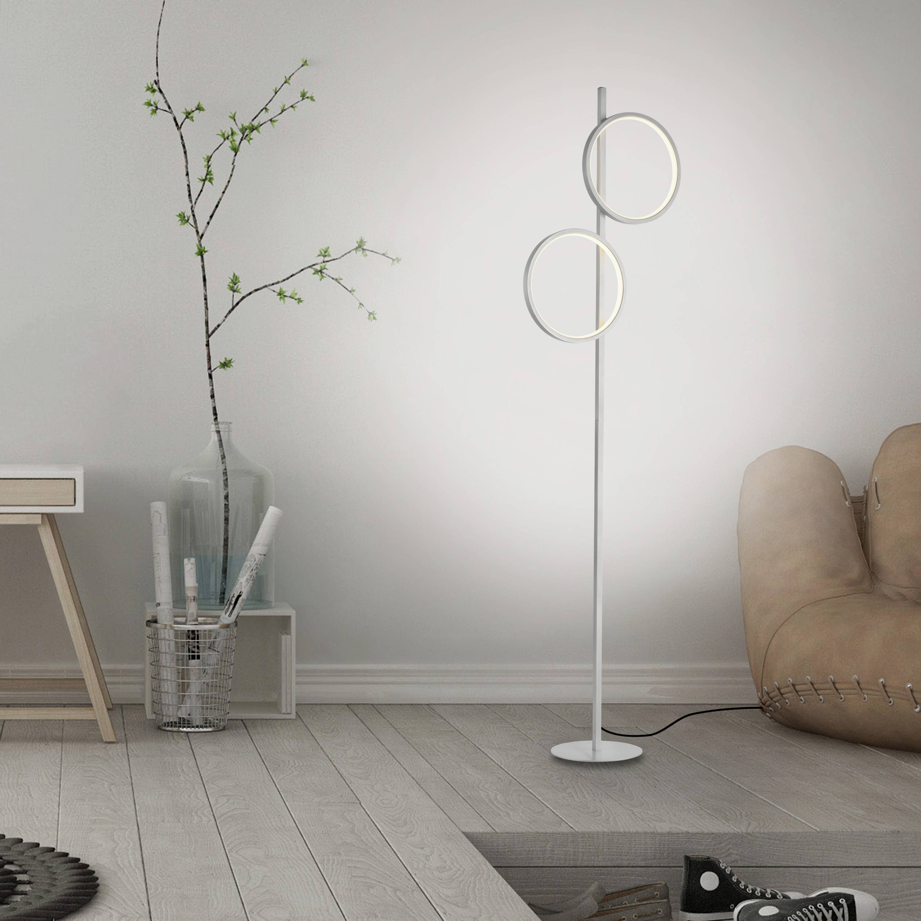 Zimmer mit moderner Stehlampe, die zwei ringförmige Leuchten umfasst. Im Hintergrund stehen Pflanzen in einer Vase und ein Ledersessel.