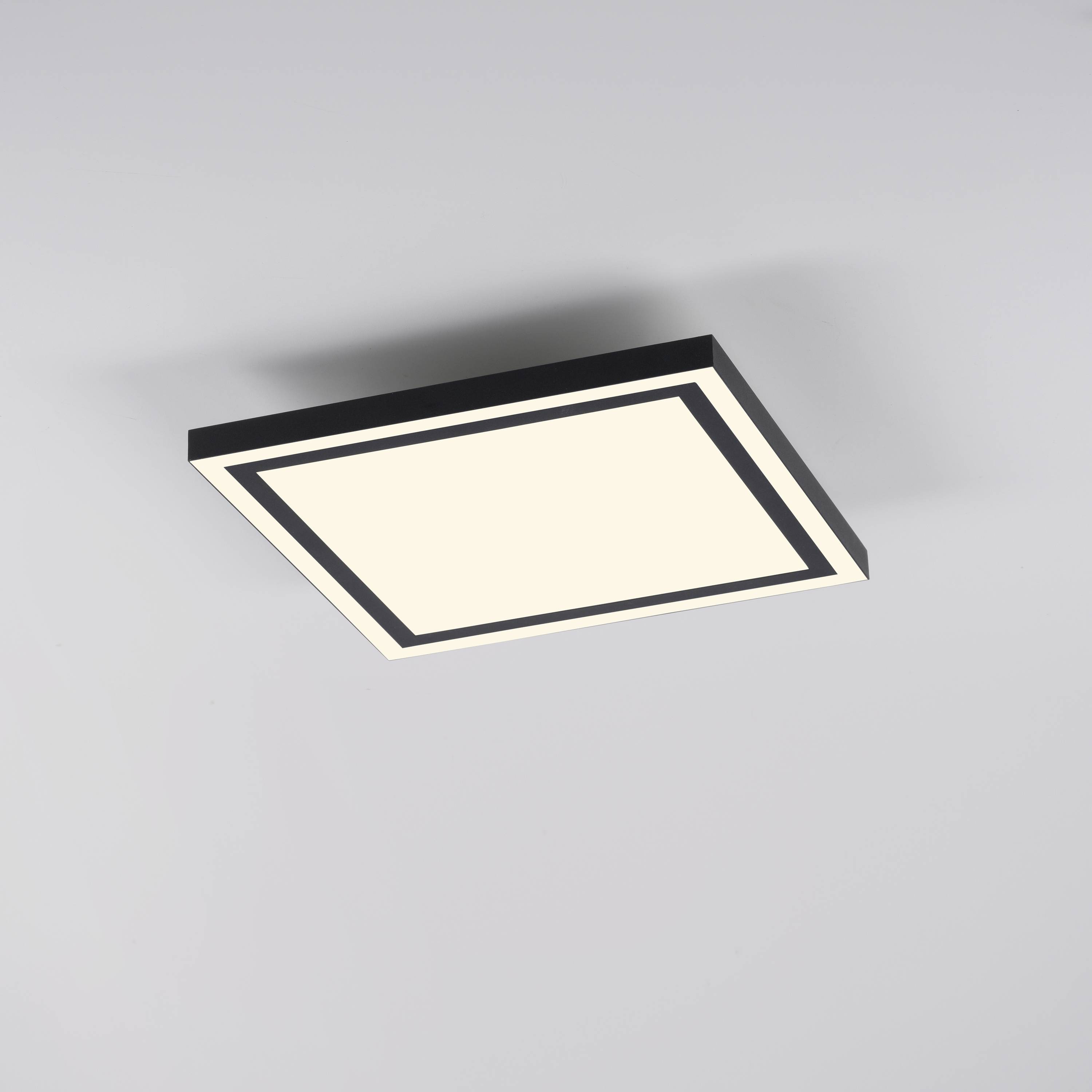 Eine quadratische Deckenleuchte mit schwarzem Rahmen und weiß leuchtender Oberfläche an einer grauen Decke montiert.