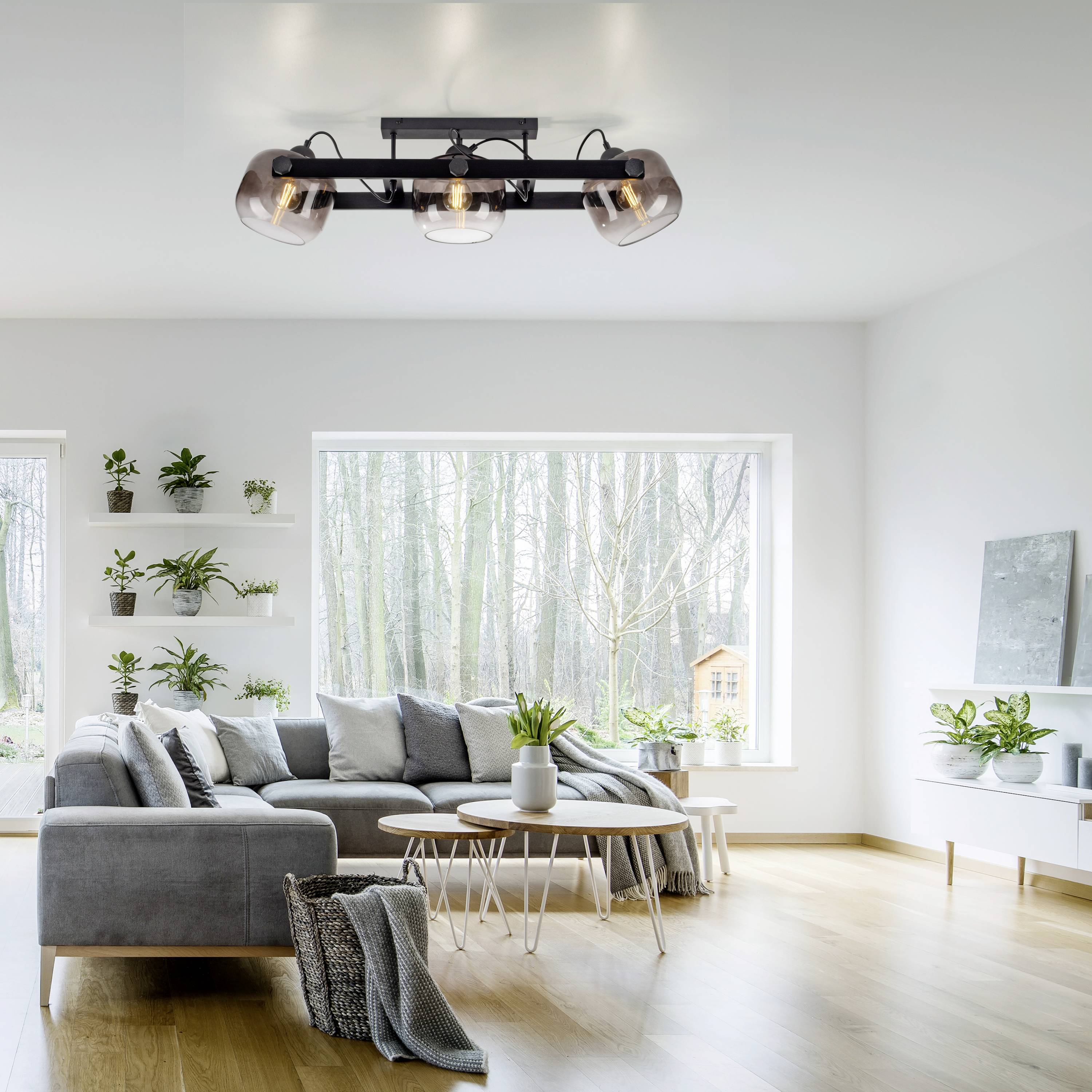 Ein helles Wohnzimmer mit moderner Deckenleuchte, grauem Sofa, Pflanzen und großem Fenster mit Blick ins Grüne.