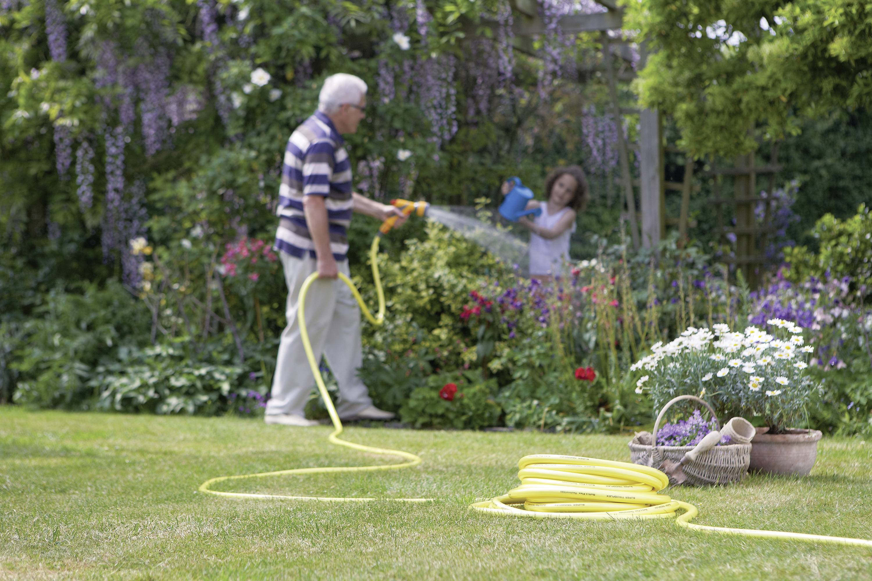 Ein älterer Mann bewässert den Garten mit einem Schlauch, während ein Kind die Blumen gießt. Im Vordergrund ein Korb mit Gartenwerkzeugen.