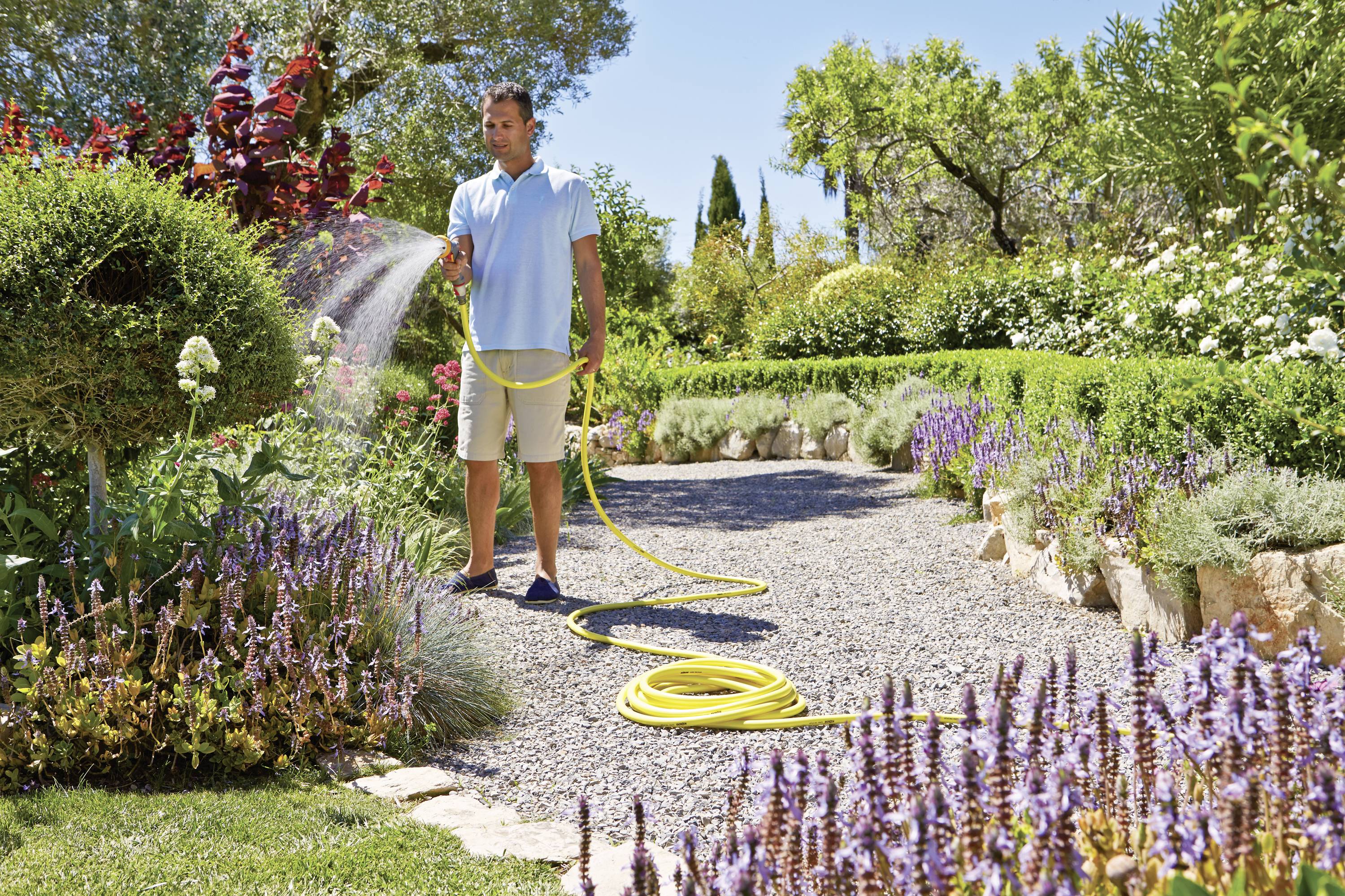 Ein Mann wässert an einem sonnigen Tag mit einem Gartenschlauch Pflanzen in einem gepflegten Garten mit blühenden Blumen und Bäumen.