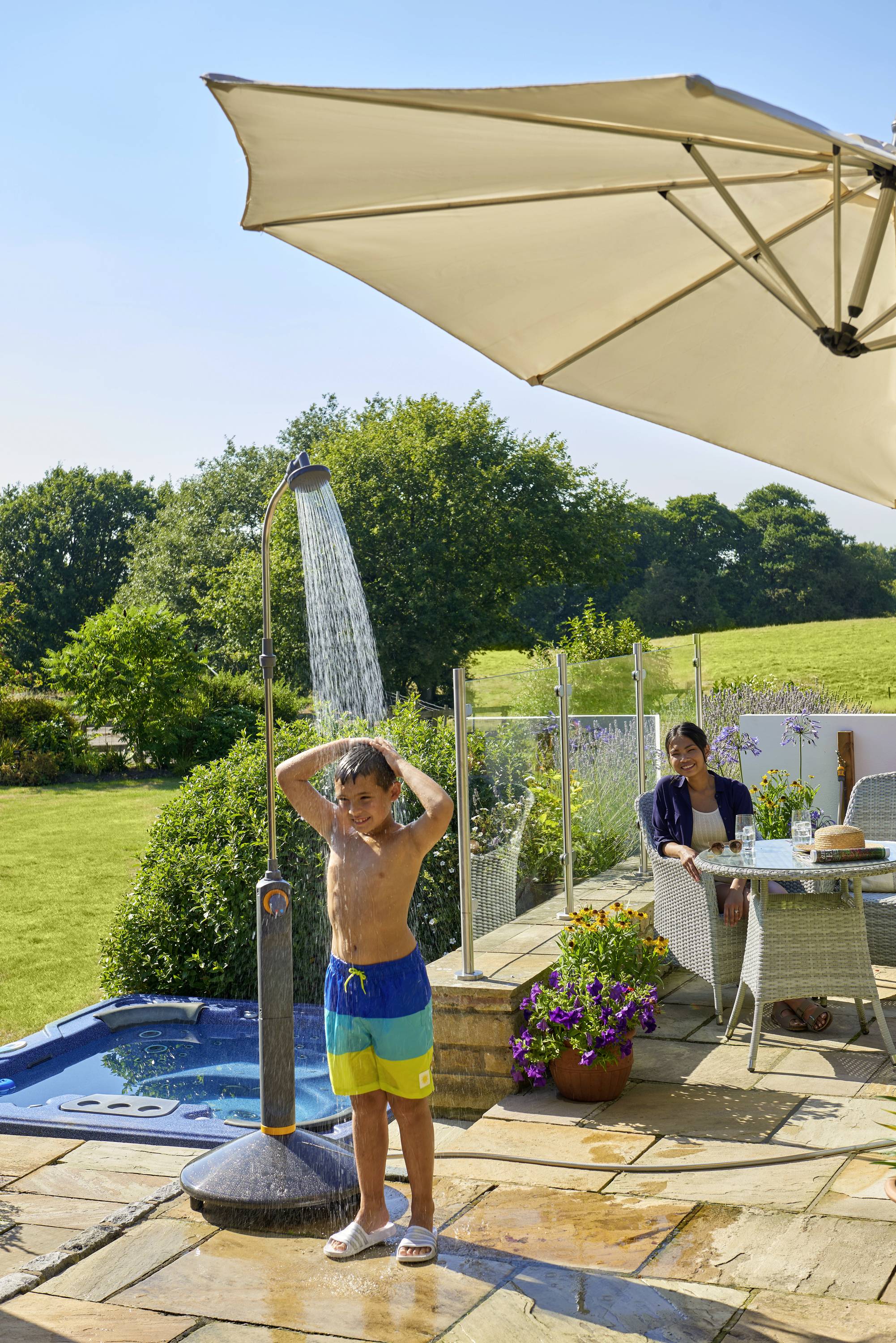 Ein Kind duscht auf einer Terrasse unter freiem Himmel, während eine sitzende Person unter einem Sonnenschirm zuschaut. Im Hintergrund ist ein Garten.