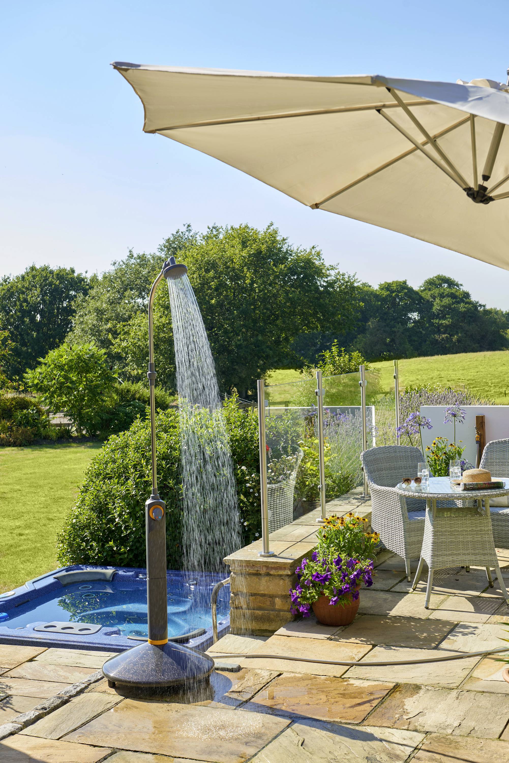 Garten mit Sonnenliege, Sonnenschirm, Tischgruppe und Gartendusche. Im Hintergrund ein Whirlpool und grüne Landschaft.