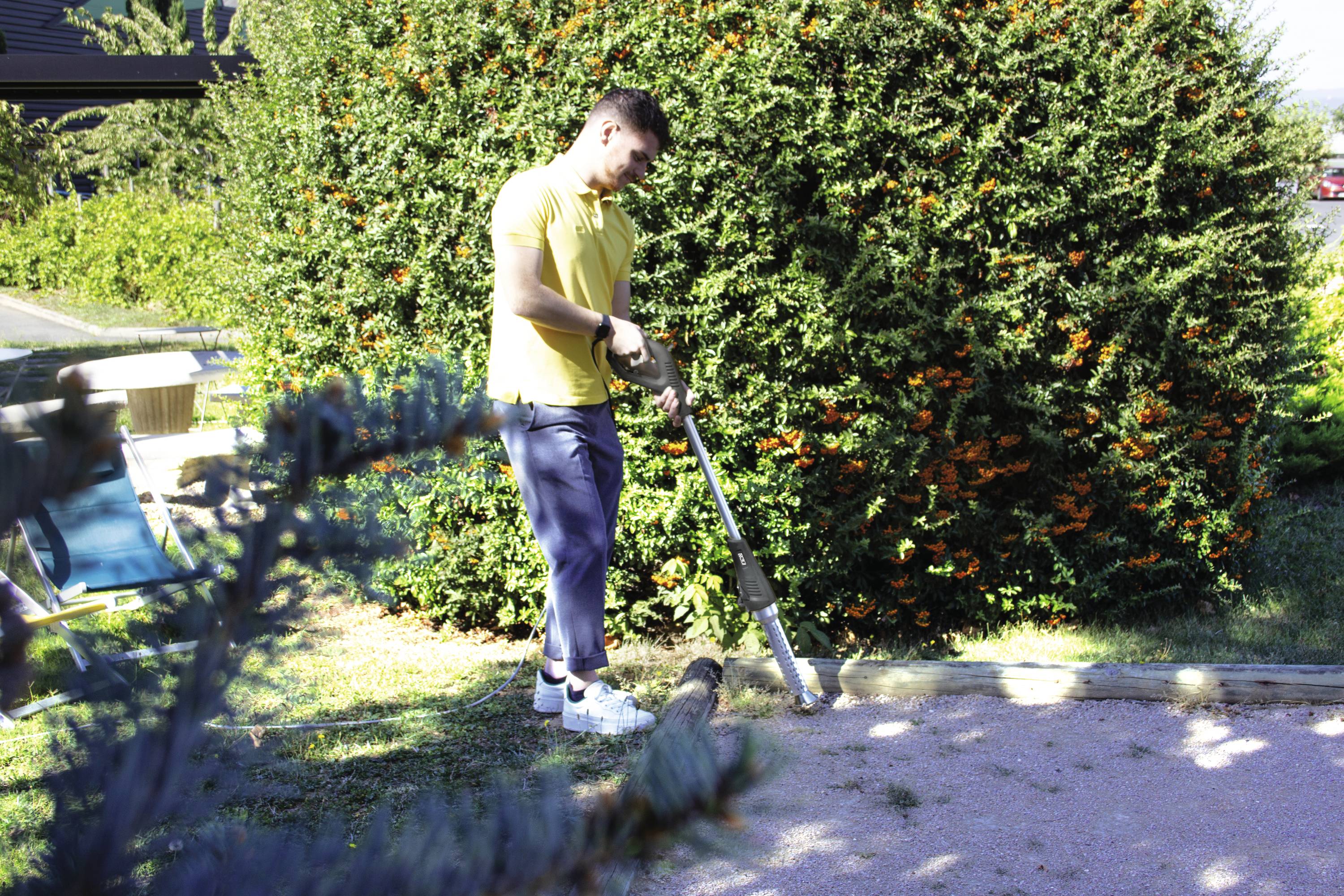 Ein Mann im gelben T-Shirt arbeitet mit einer Gartenmaschine auf einem Rasen neben einem großen Busch mit orangefarbenen Beeren.