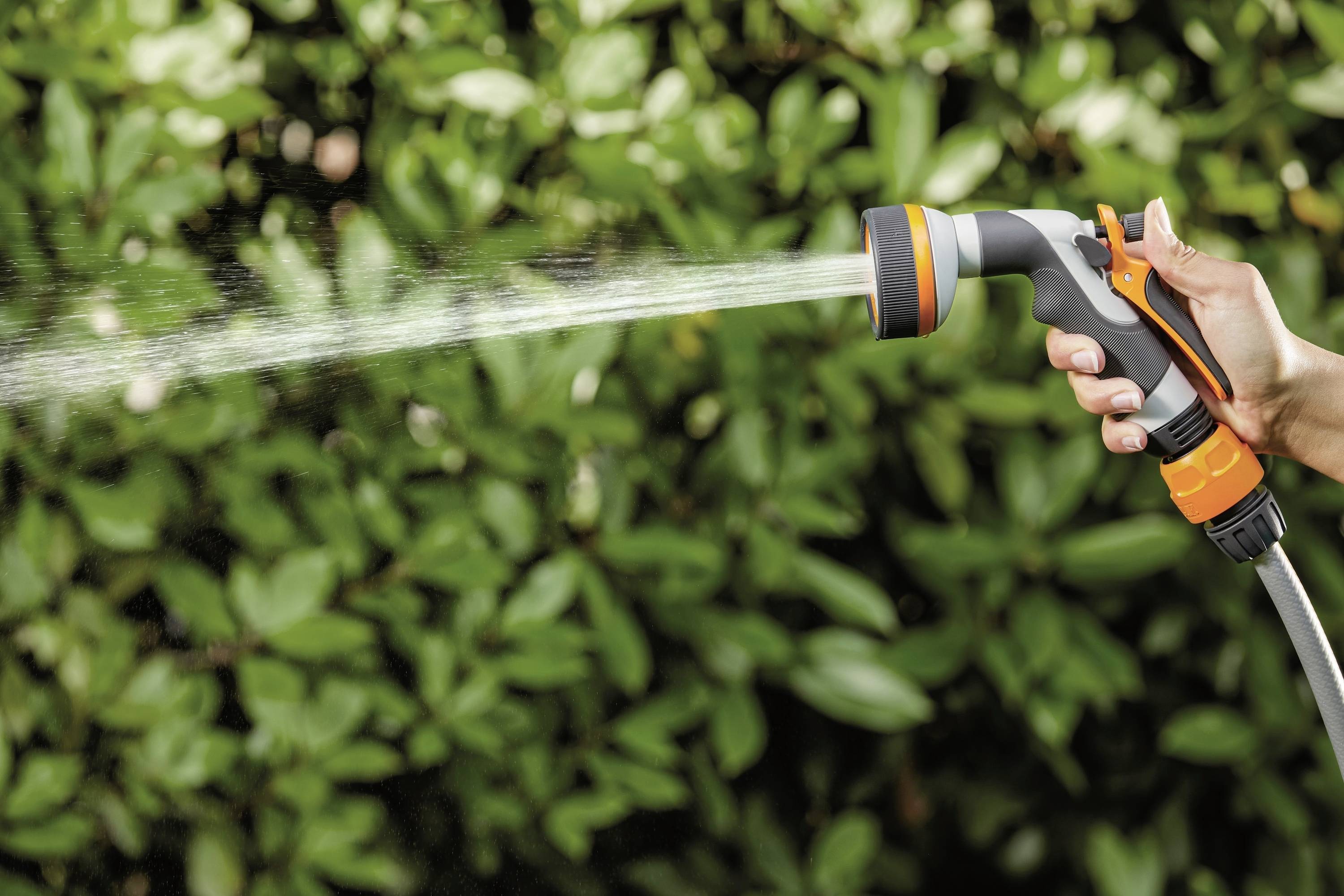 Hand spritzt mit einer Gartenbrause Wasser auf grüne Büsche im Hintergrund, zeigt Gartenpflege oder Bewässerungssituation.