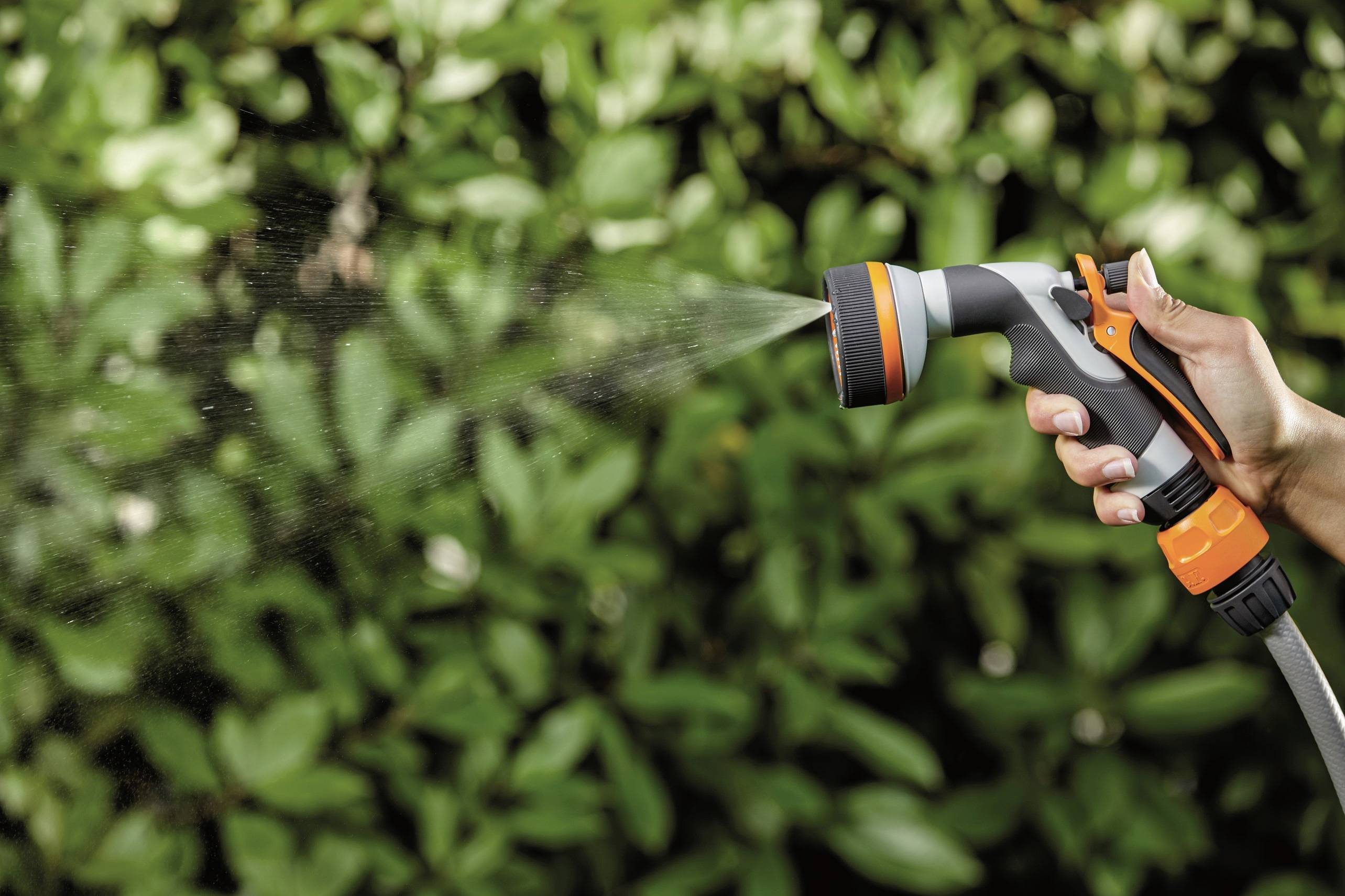 Eine Hand hält eine Gartenspritzdüse, die Wasser auf grüne Blätter sprüht. Hintergrund unscharf mit dichtem, grünem Laub.