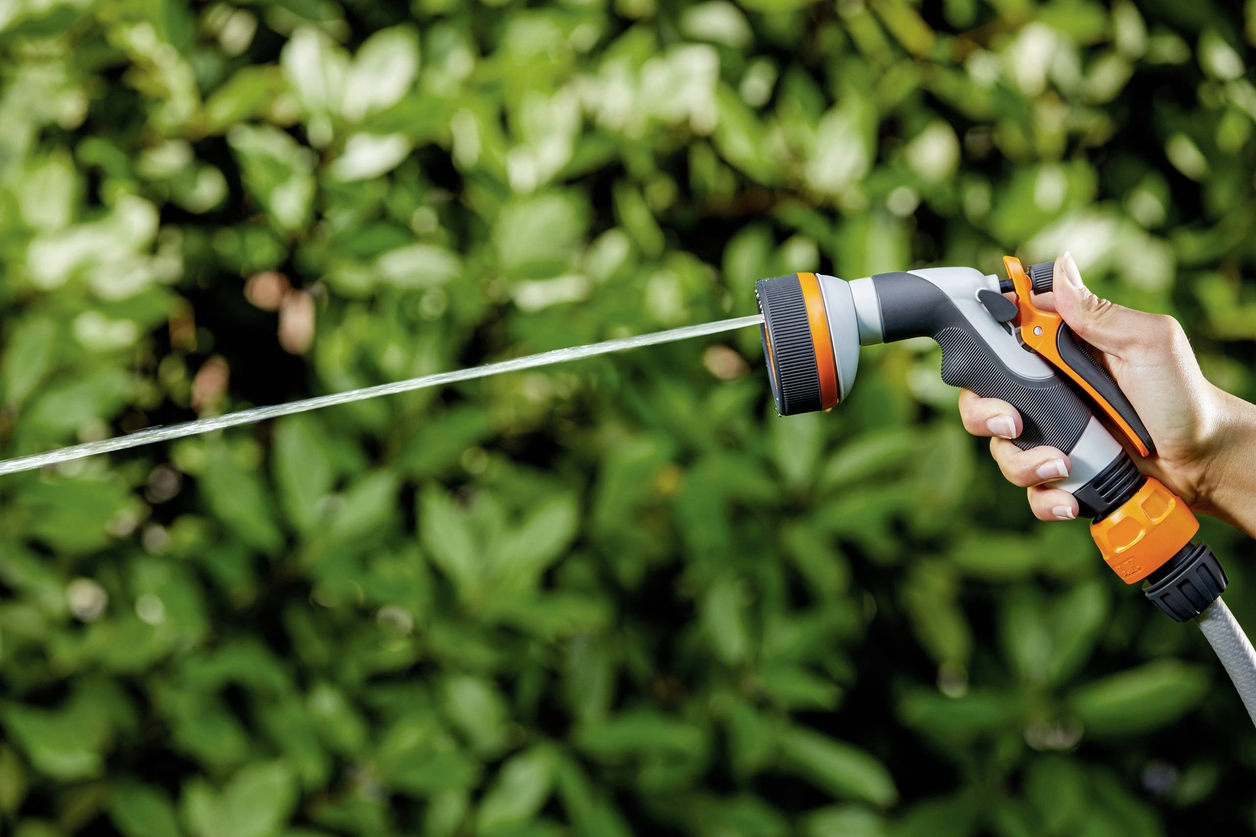 Eine Hand hält eine Gartenspritze, aus der ein Wasserstrahl auf grüne Blätter im Hintergrund spritzt.