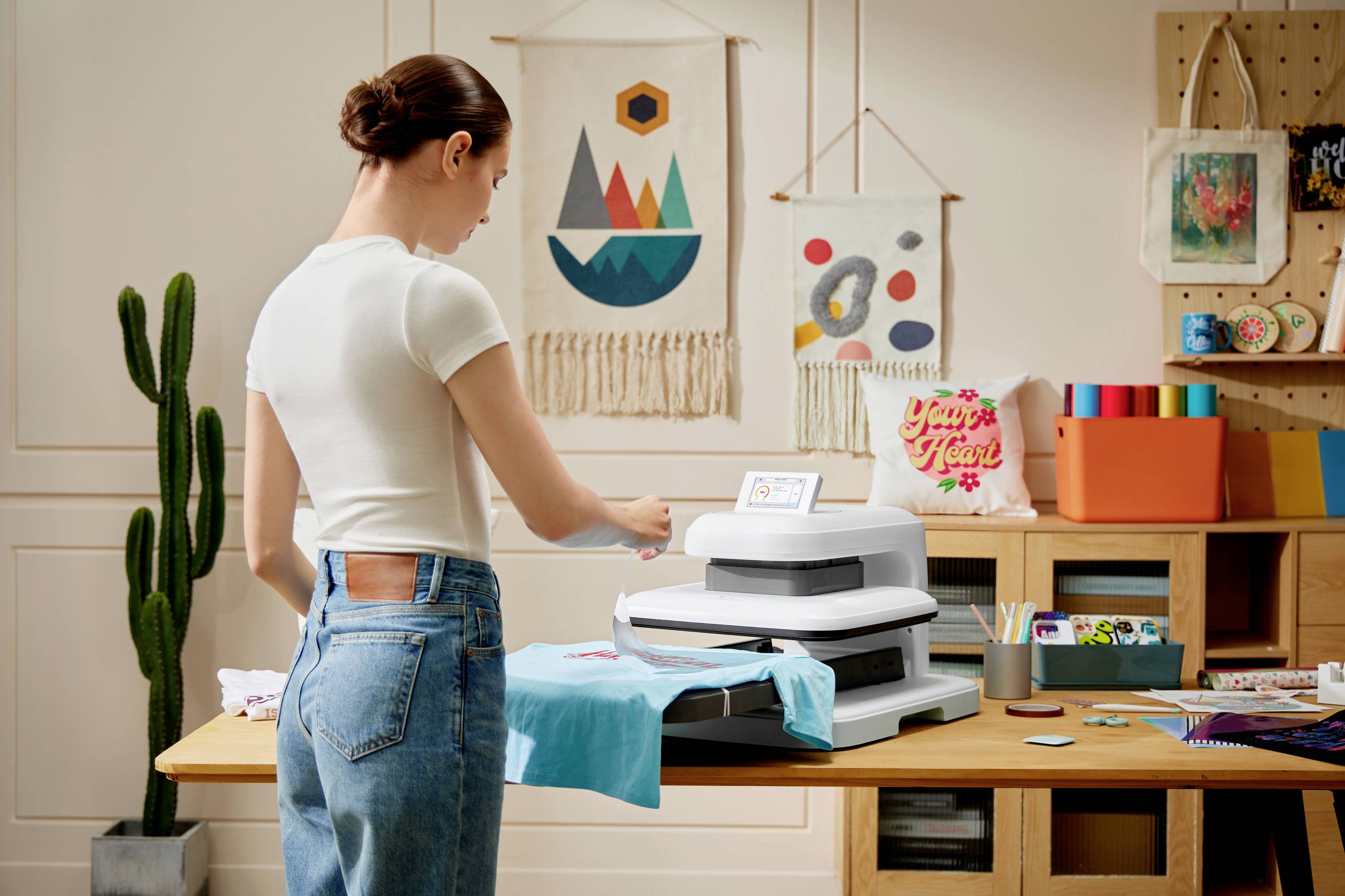 Eine Person bedruckt ein türkisfarbenes T-Shirt mit einem Drucker auf einem Holztisch. Wanddekorationen und Pflanzen im Hintergrund.