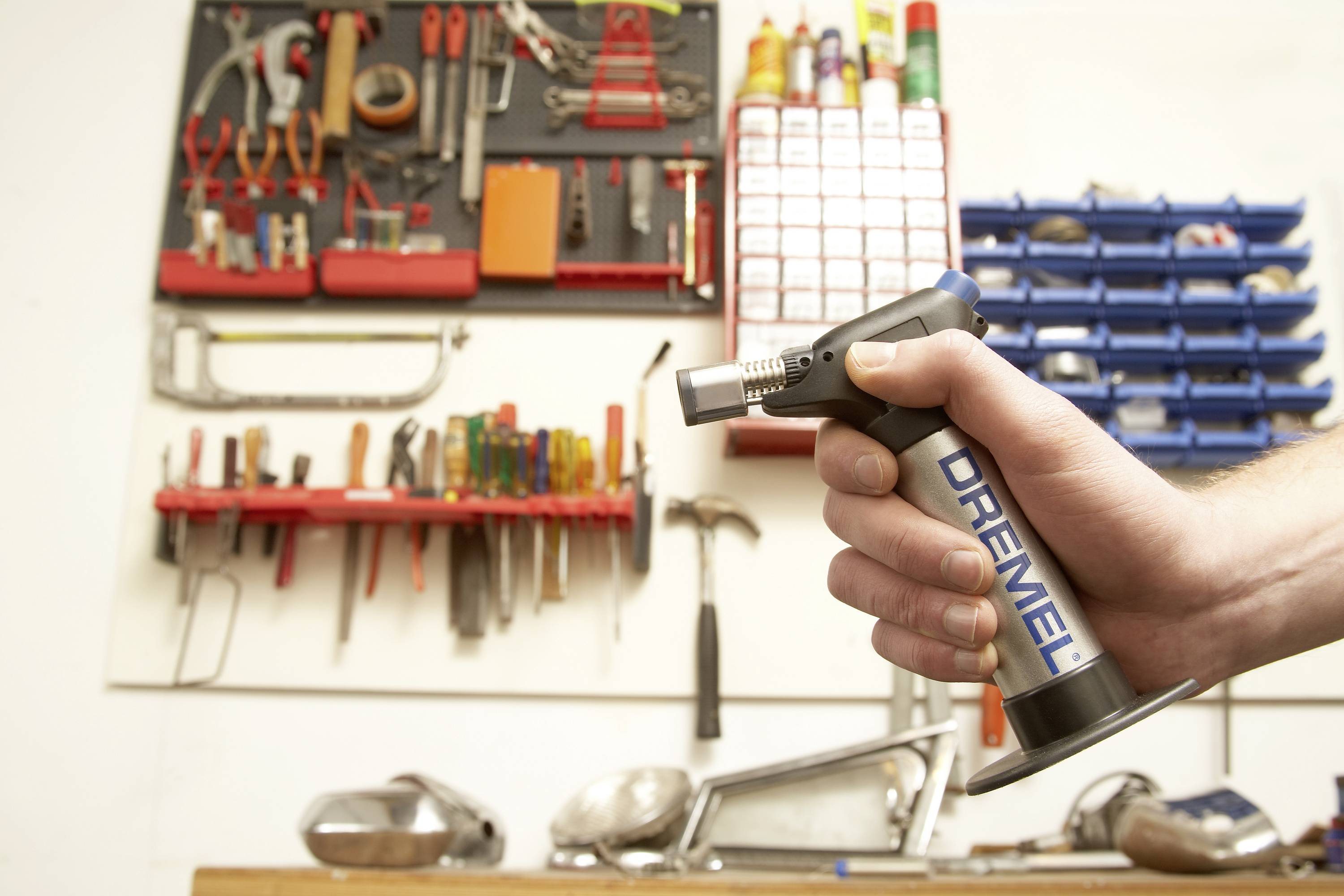 Eine Hand hält einen Dremel-Lötkolben in einer Werkstatt mit Haken an der Wand, auf denen Werkzeuge und Materialien organisiert sind.